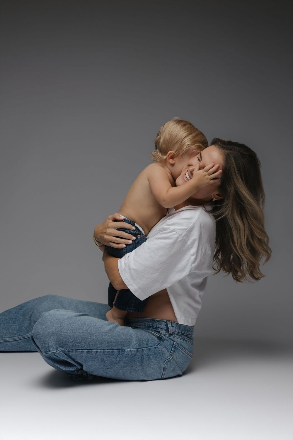 A mother in white crop top and jeans sits on floor hugging her toddler child in a warm studio embrace.