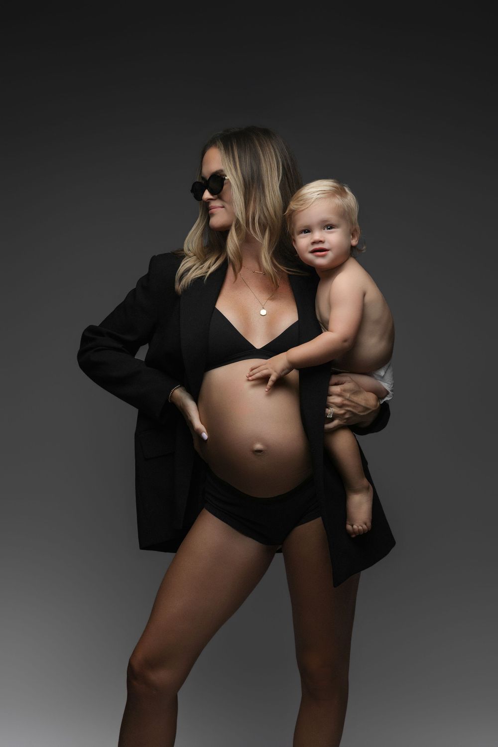 Pregnant woman in black bikini and blazer holding a toddler, posing confidently in a studio setting.