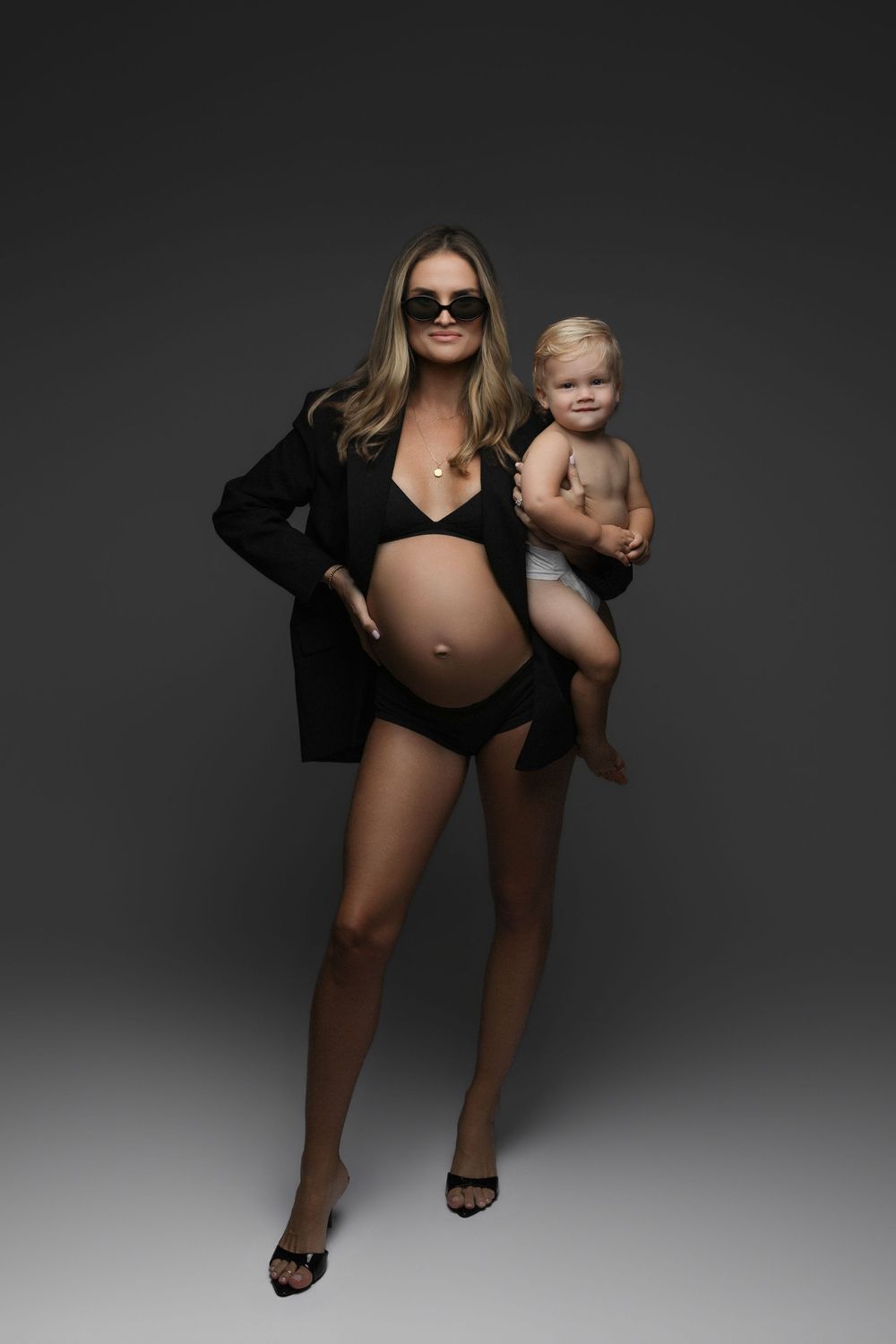Pregnant woman in black bikini and blazer holding a baby, wearing sunglasses and heels on a dark background.