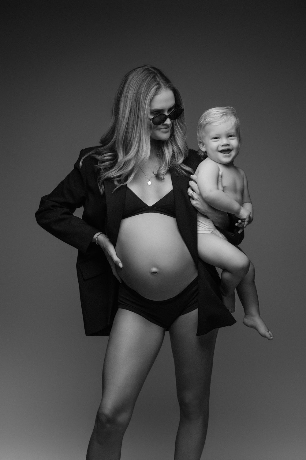 Black and white photo of a pregnant woman in bikini and blazer holding a smiling toddler in a studio setting.