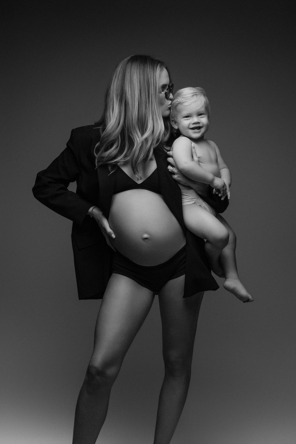Black and white photo of a pregnant woman in a blazer holding a smiling baby, showcasing maternity beauty.