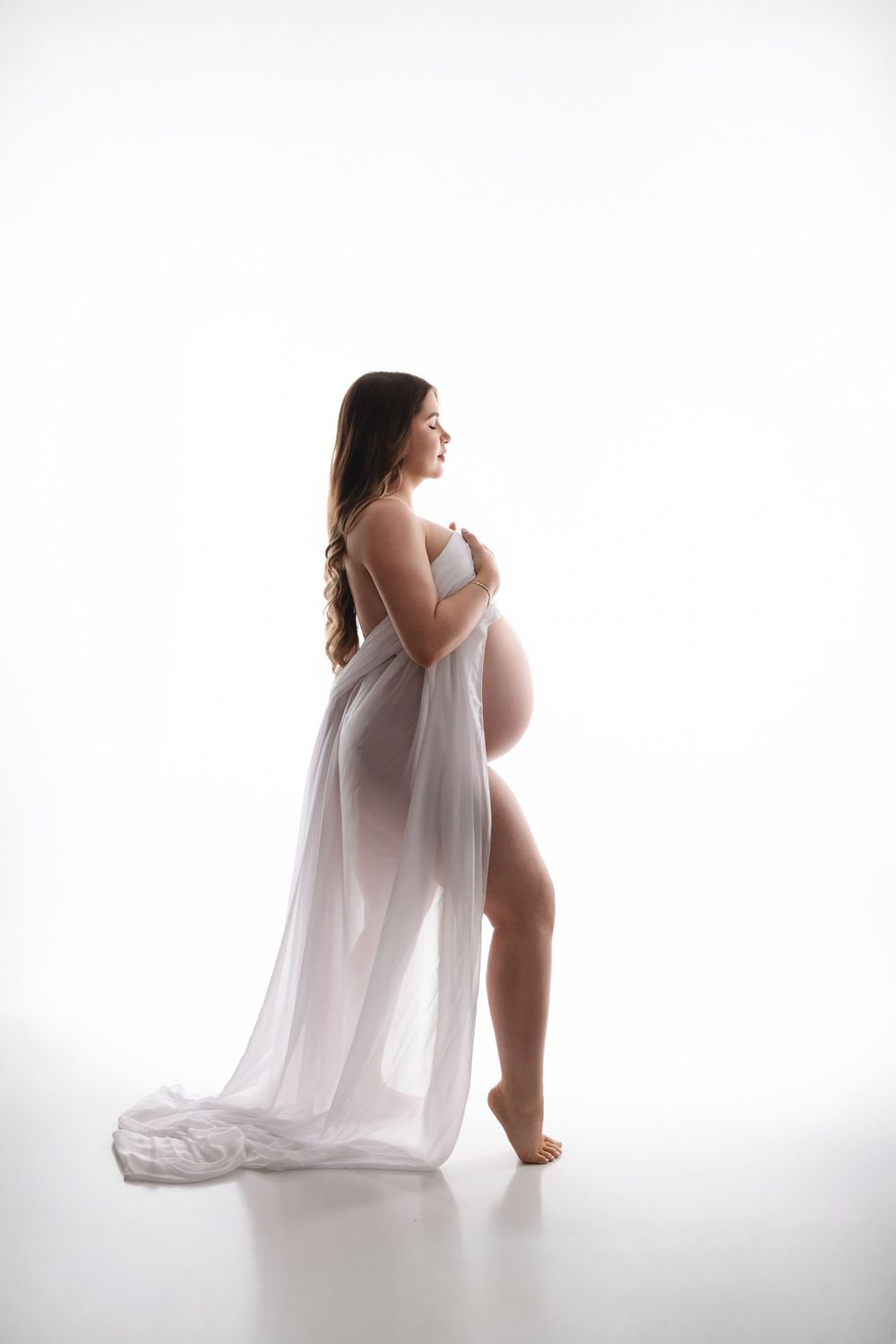 Pregnant woman in sheer white fabric draped elegantly, posing sideways in a bright studio for a maternity photo shoot.