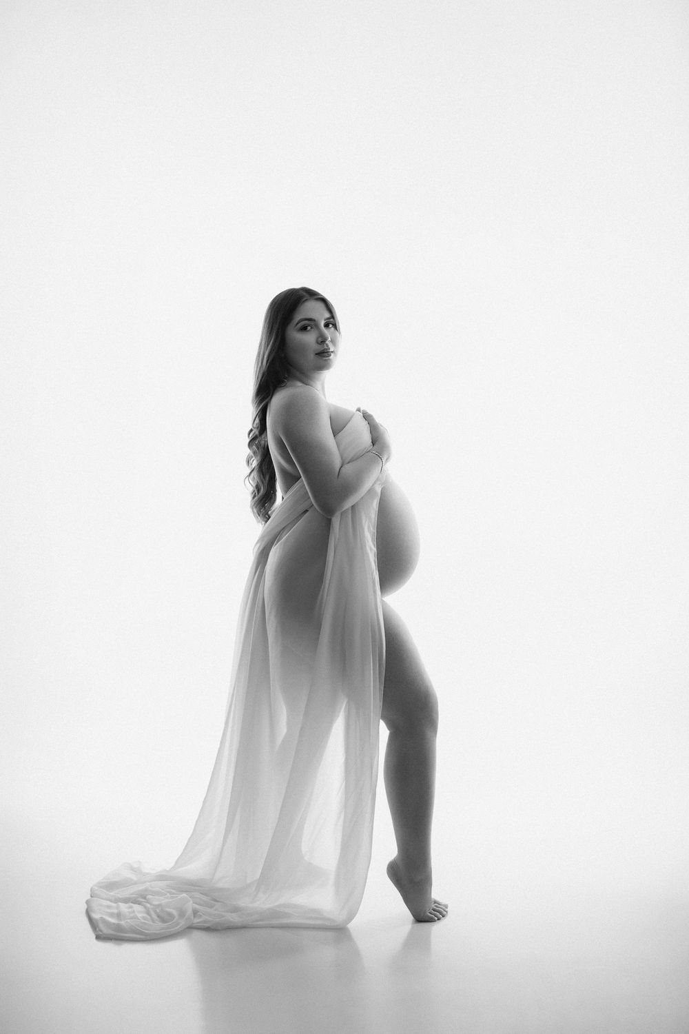 Black and white maternity photo of a pregnant woman draped in sheer white fabric, posing elegantly in a studio.