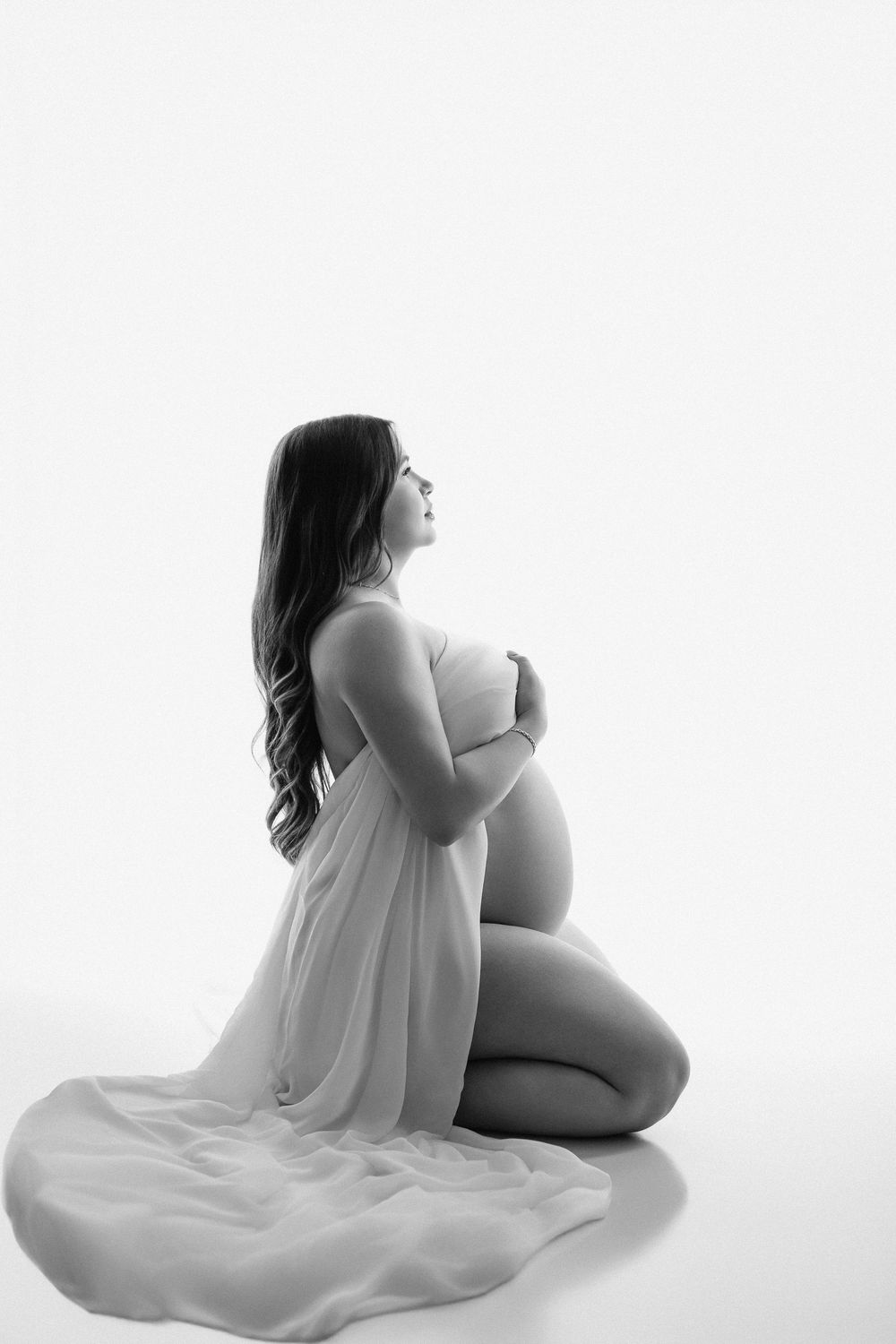 Black and white maternity photo of pregnant woman kneeling in sheer fabric, cradling her belly against white background.