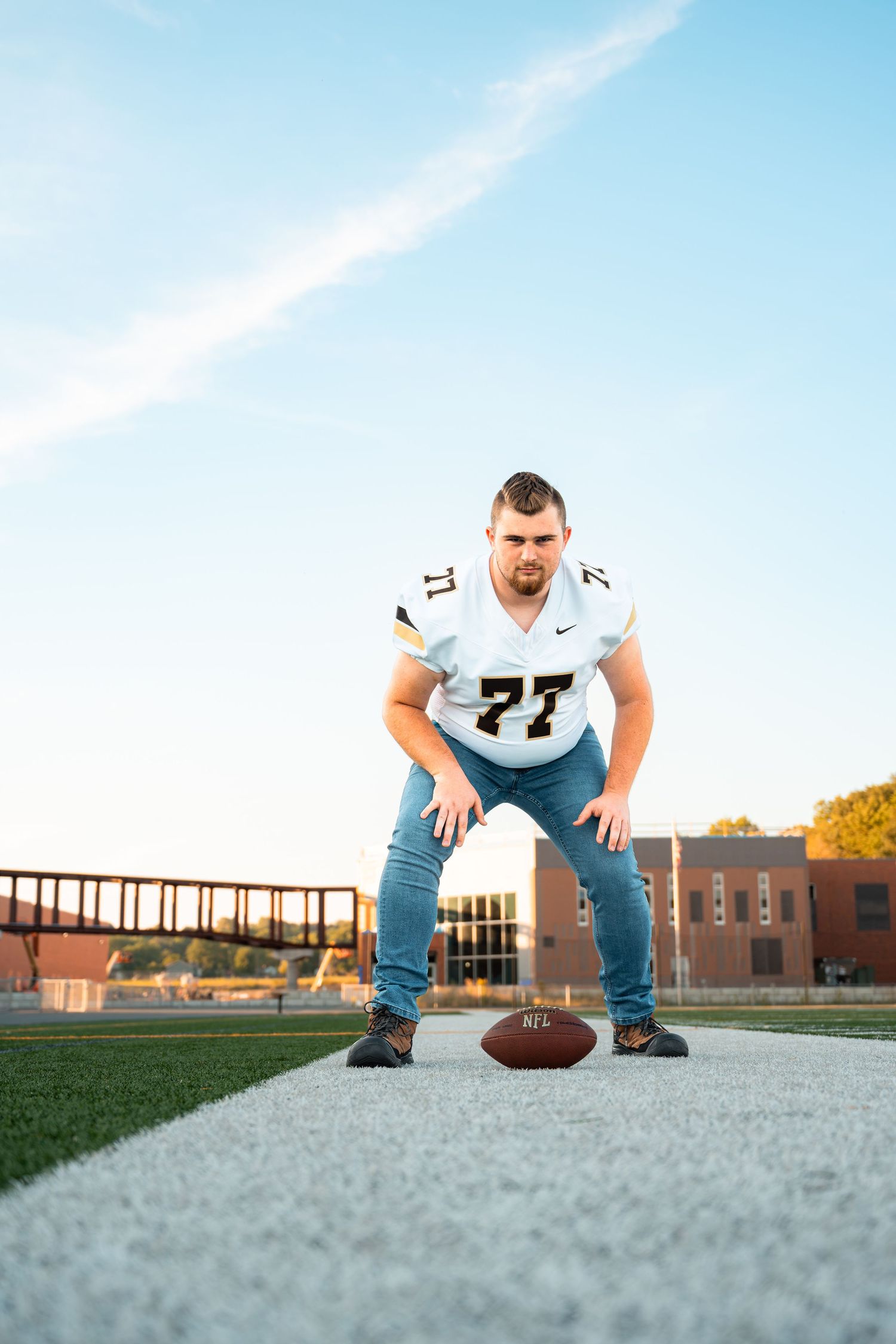 Football player in white football jersey crouches over ball on green field with school building backdrop.