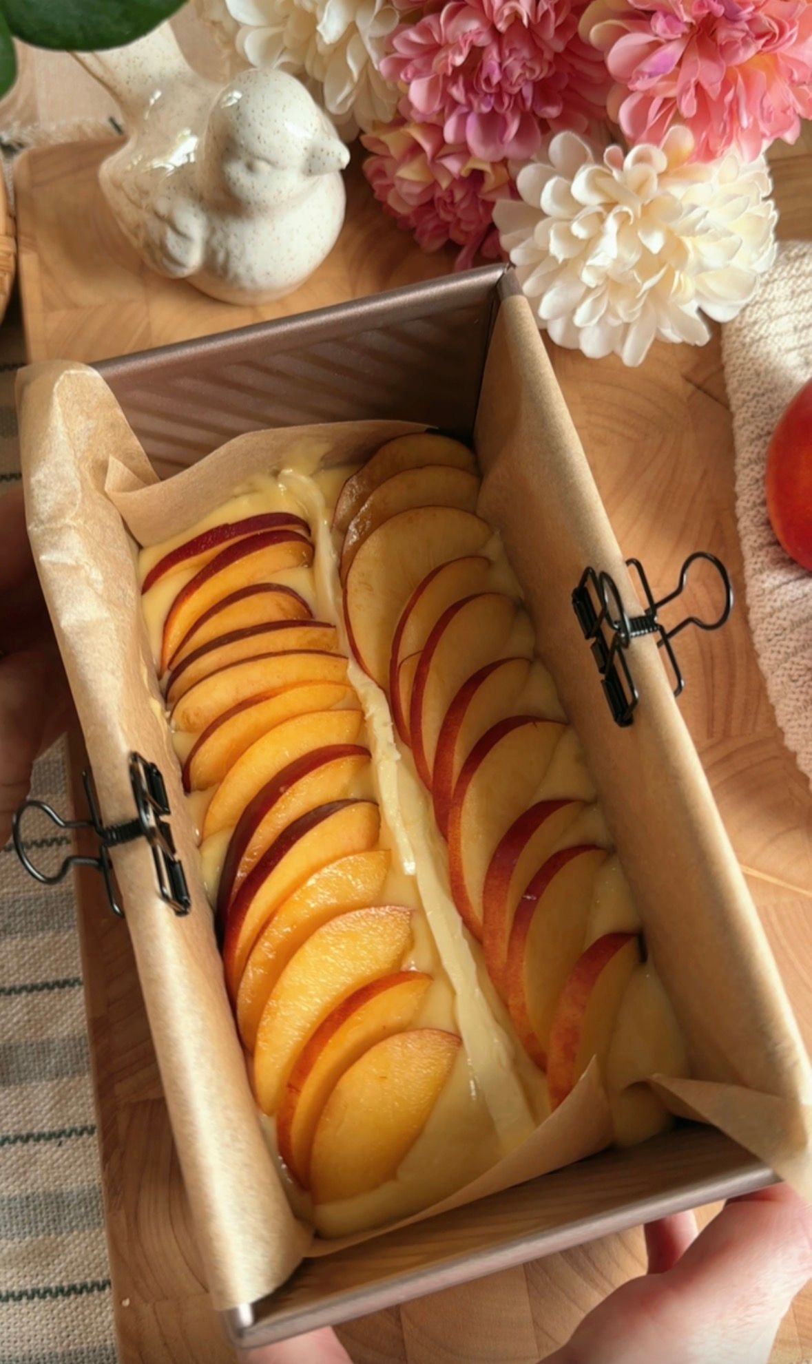 Golden peach loaf cake in parchment-lined pan, fresh peaches and pink flowers on wooden board.