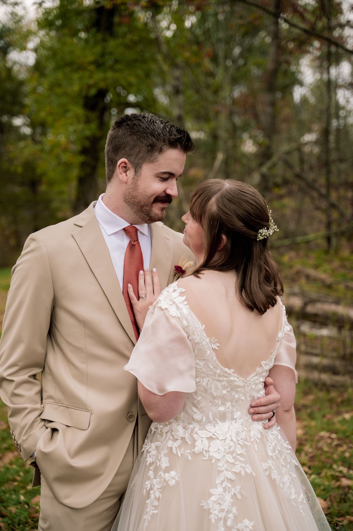 Bride and groom share a romantic moment outdoors in fall, she in lace gown, he in tan suit with coral tie.