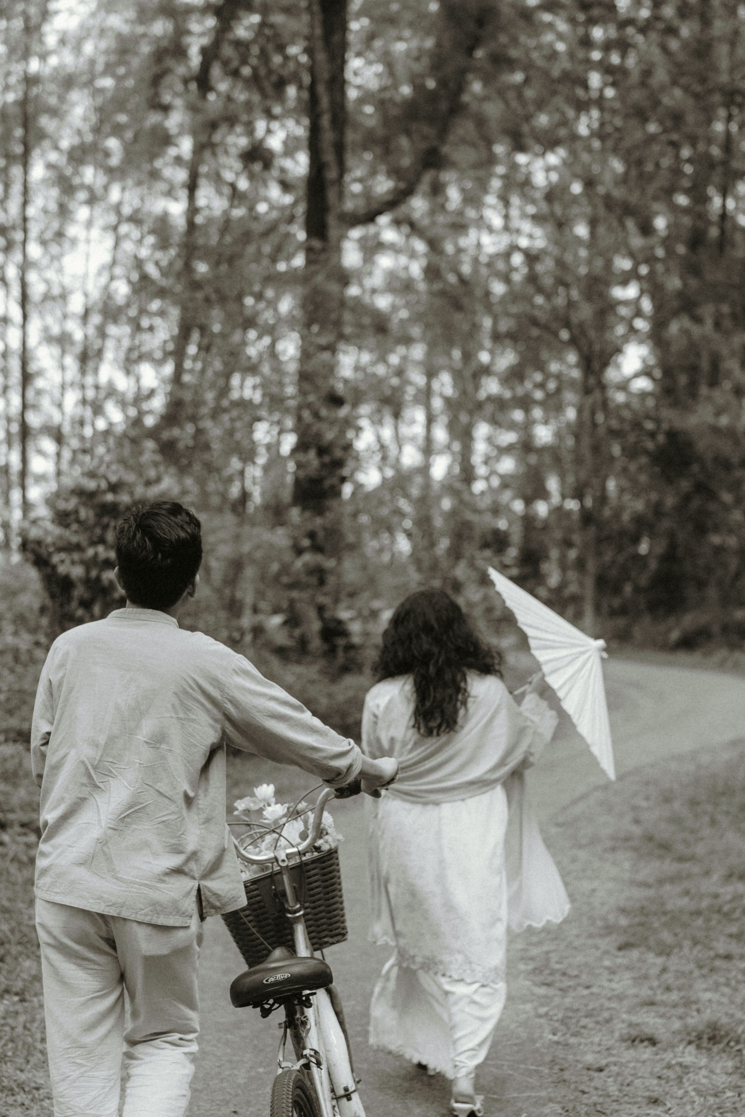 Couple walking through forest path with bicycle and parasol in black and white photo.