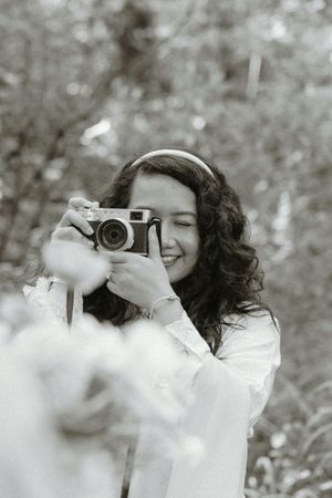 Young woman smiling while holding a vintage camera outdoors, captured in black and white photography.