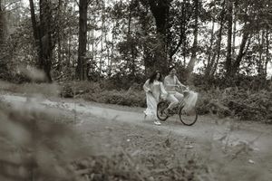 Couple riding a bicycle together on a dirt path surrounded by lush trees in a black and white photo.