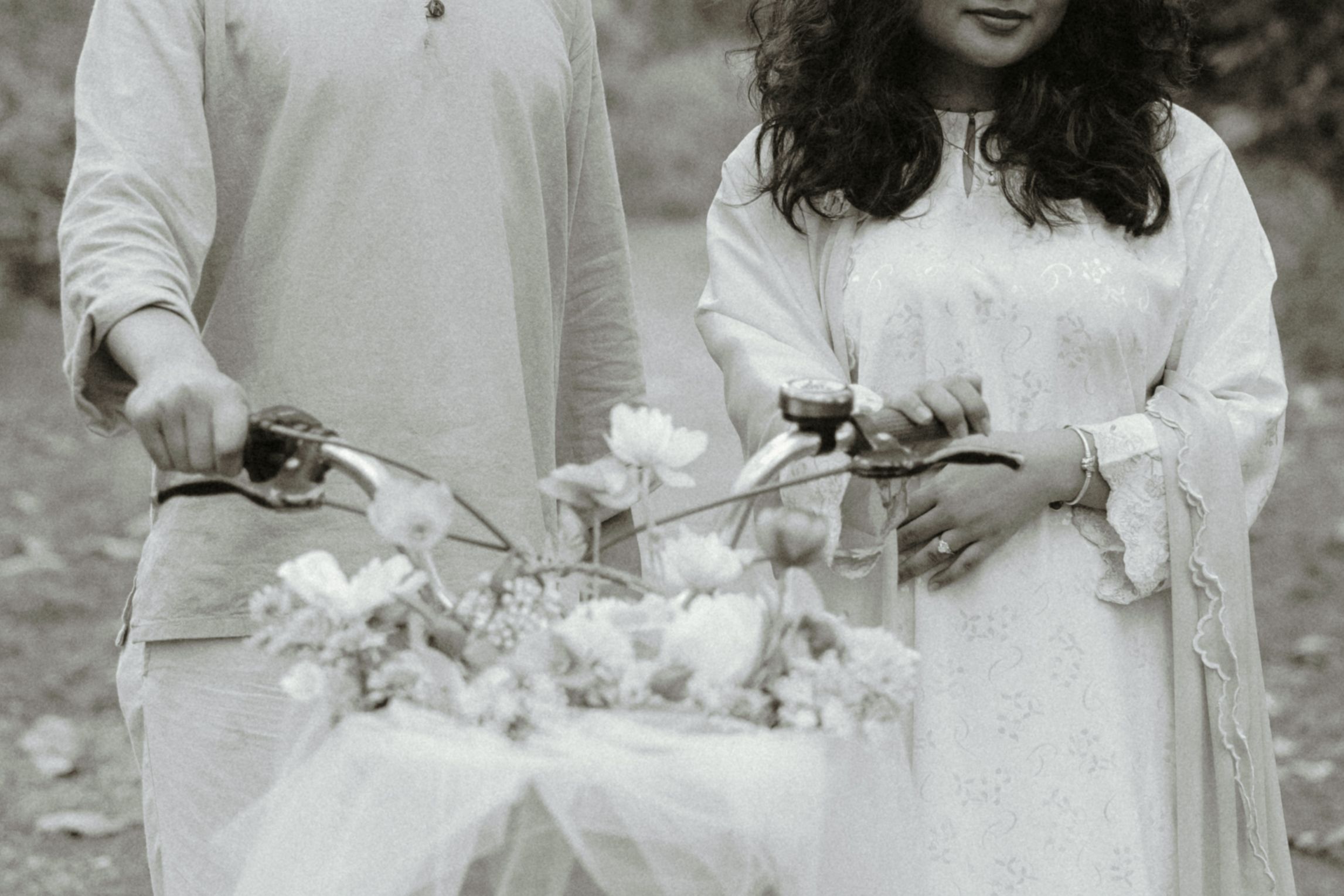 Black and white photo of a couple in white traditional attire holding a bicycle adorned with flowers.