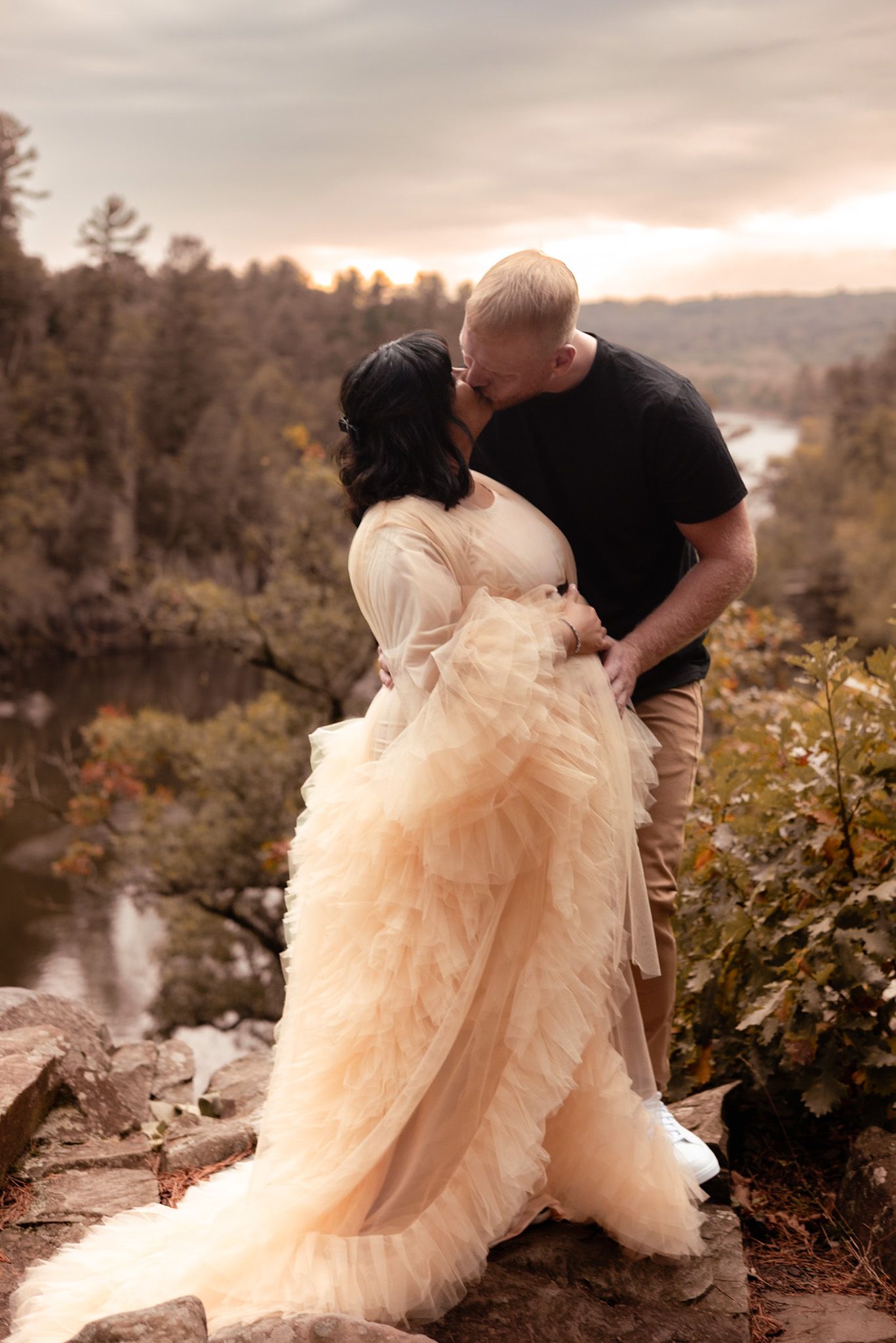 Couple kissing in a romantic outdoor setting, woman in a flowing champagne tulle gown, forest and river backdrop at sunset.