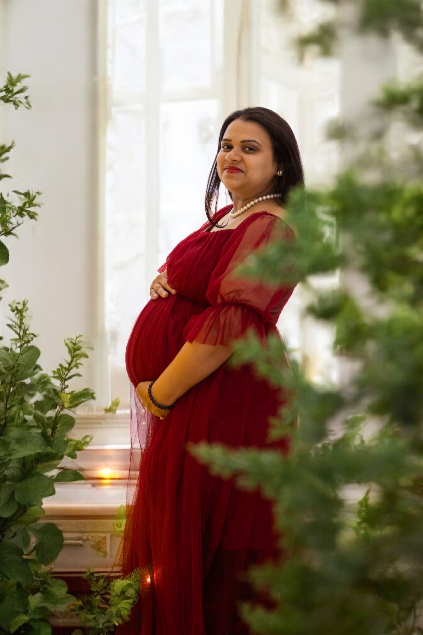 Pregnant woman in red tulle gown cradling baby bump, pearl necklace, standing by window surrounded by lush greenery.