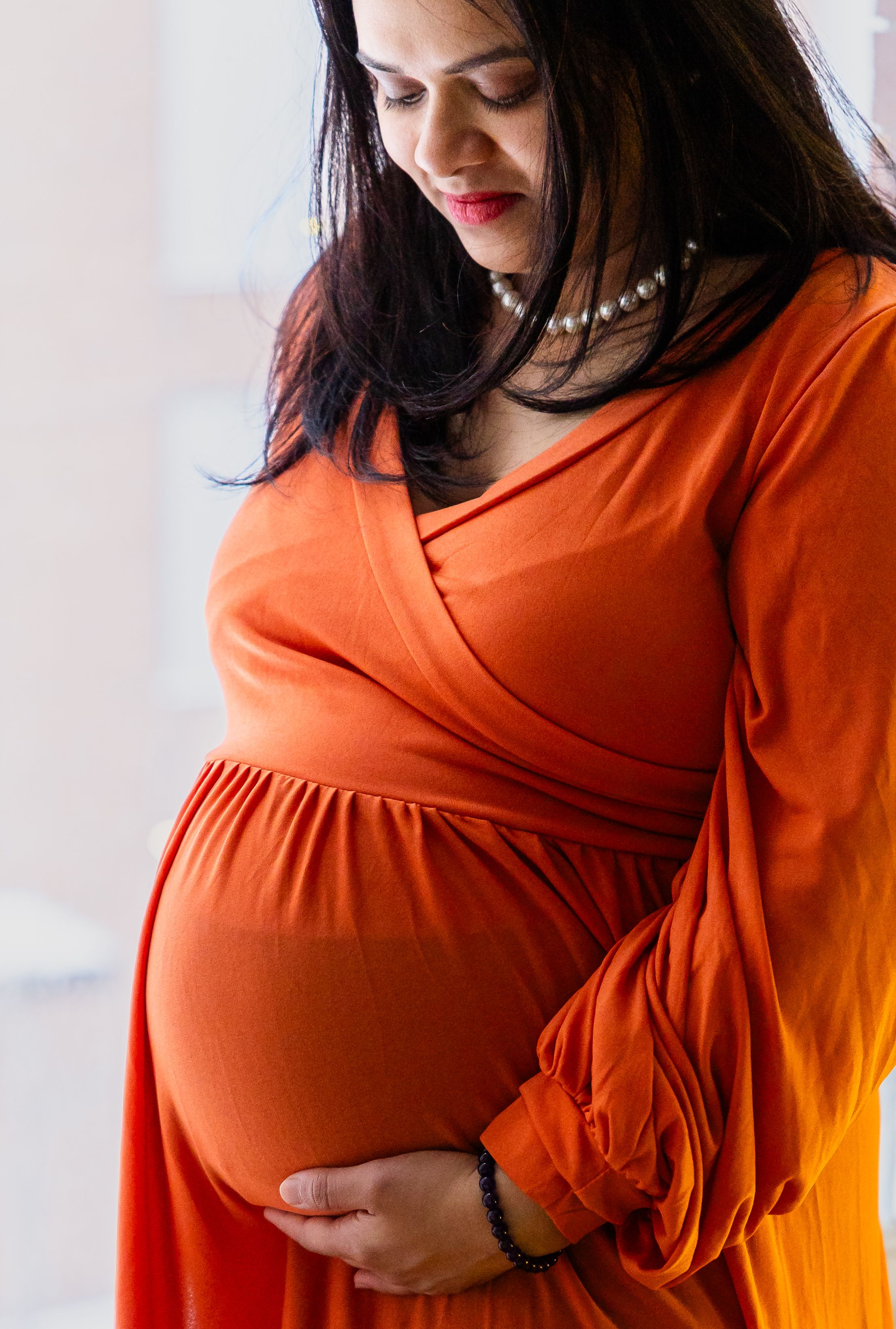 Pregnant woman in orange wrap dress cradling baby bump, wearing pearl necklace, looking down lovingly.