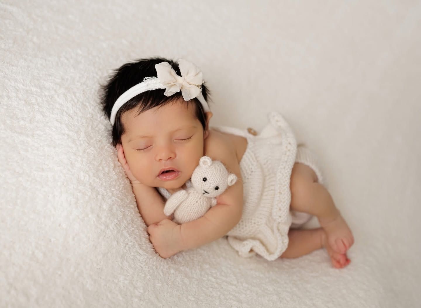 Smiling newborn baby girl in lavender lace outfit and floral headband sleeping peacefully on white backdrop.