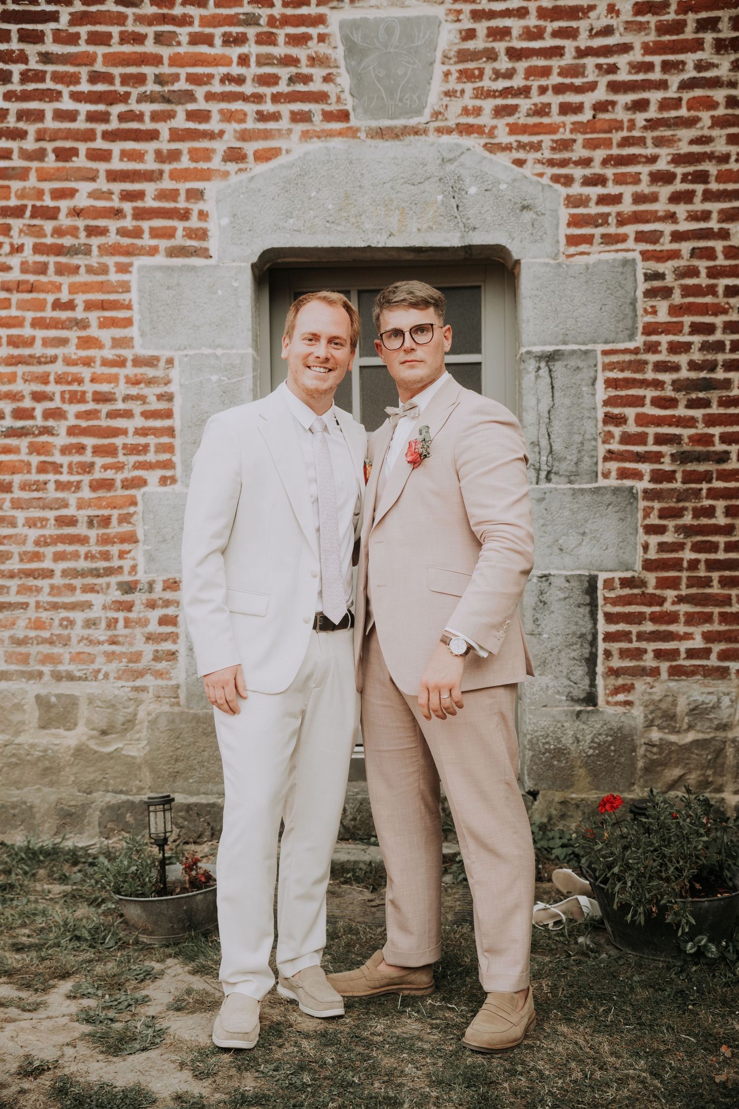 Two grooms in elegant suits pose together in front of a rustic brick building on their wedding day.
