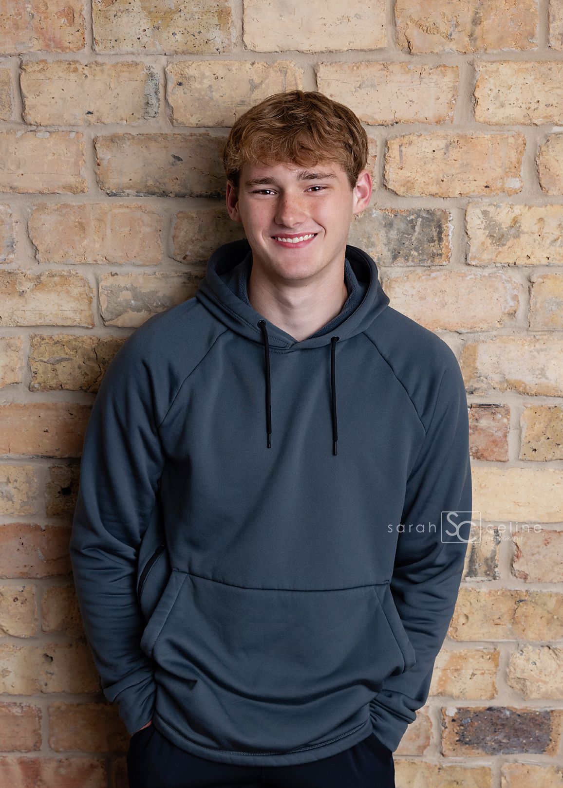 Smiling teenage boy with red hair wearing a gray hoodie, leaning against a brick wall.