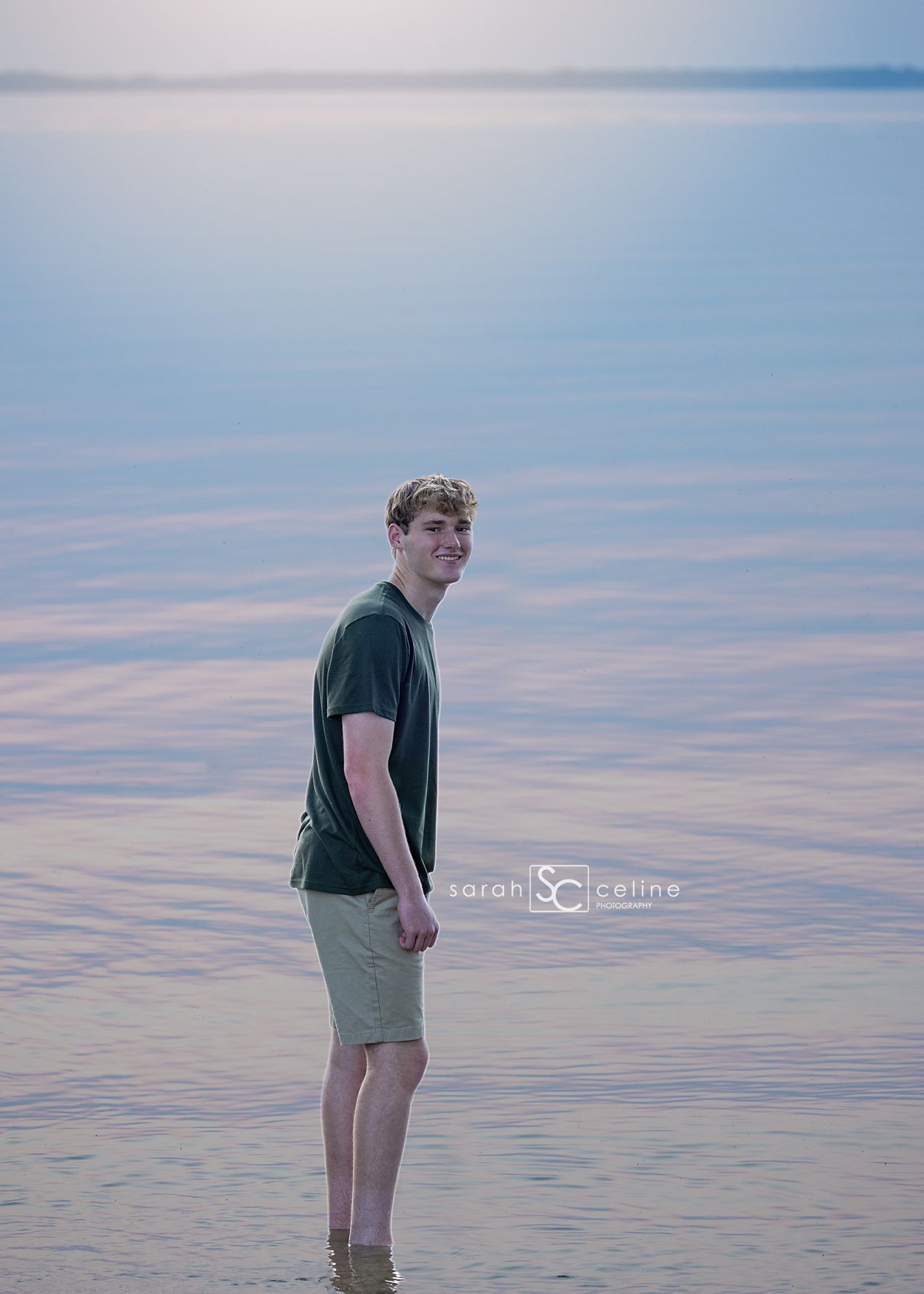 Young man smiling in shallow water at sunset, wearing green shirt and khaki shorts, professional portrait photography.
