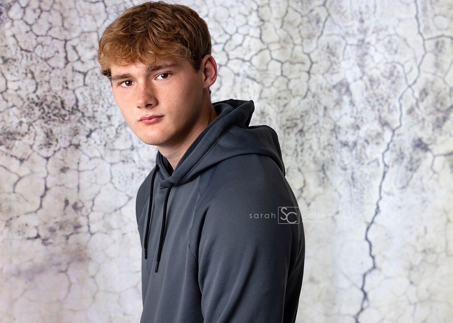 Teen boy in gray hoodie poses against cracked white backdrop in professional portrait photo.