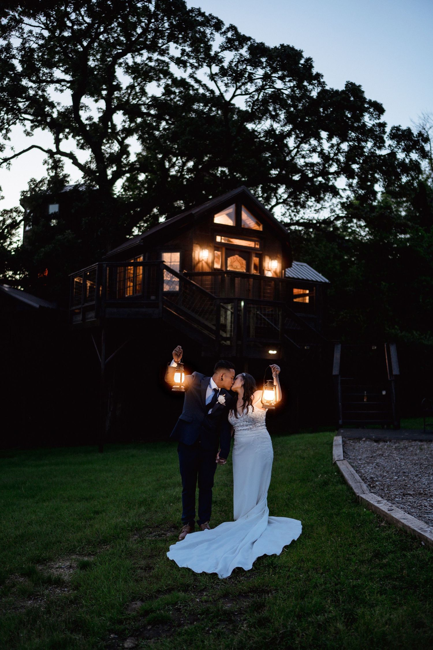 Bride and groom kissing at dusk holding lanterns in front of a warmly lit rustic treehouse venue.