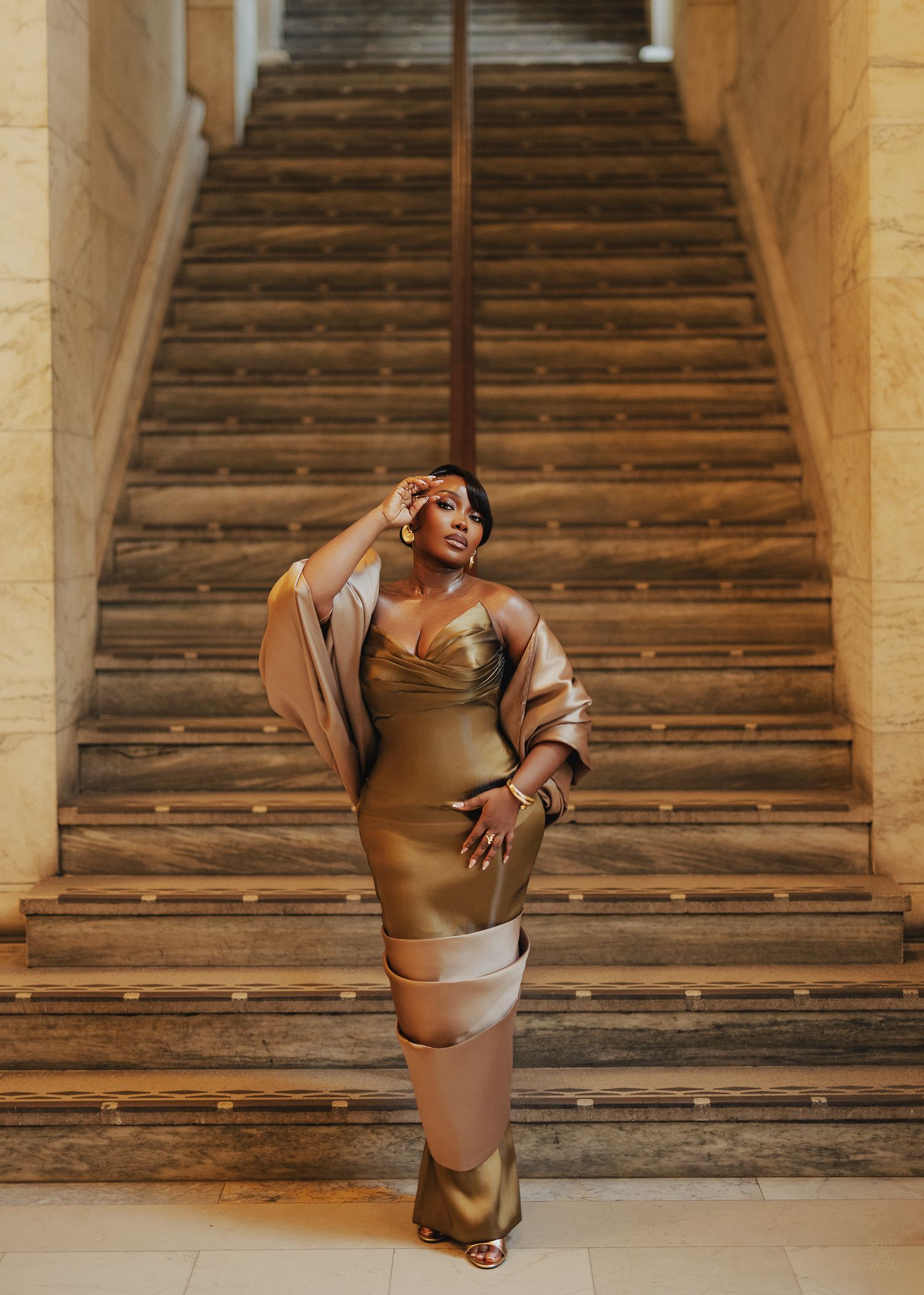 Woman in gold satin midi dress and wrap poses elegantly on marble staircase in luxurious interior setting.