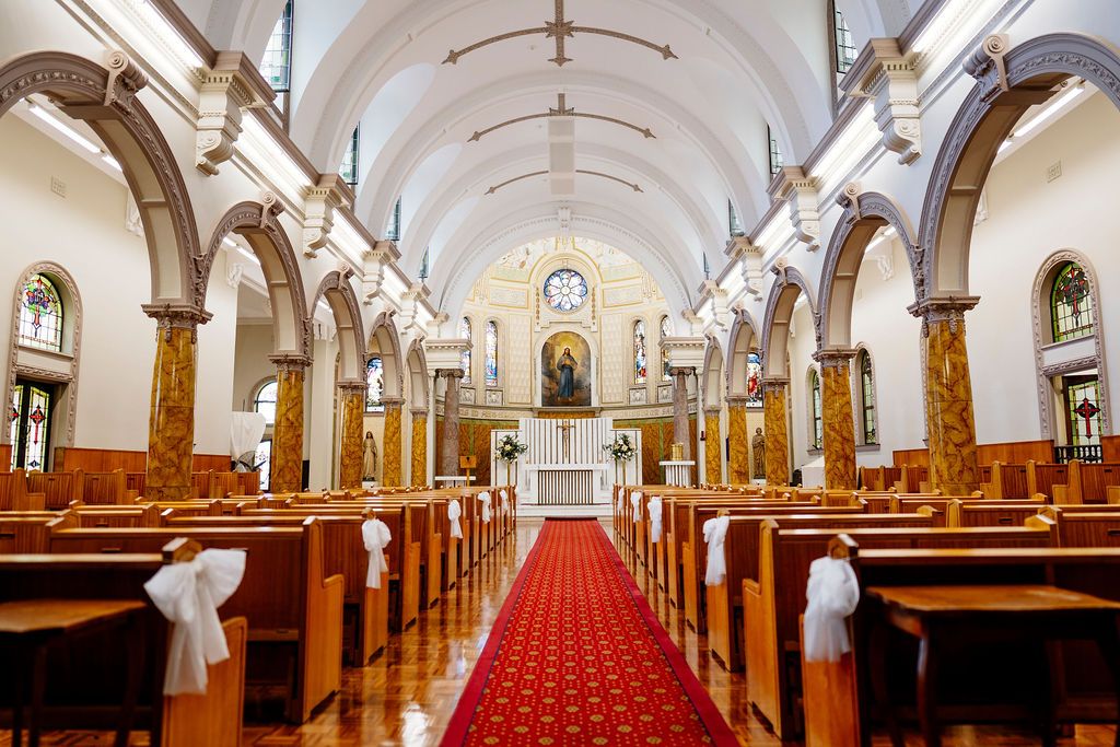 Elegant church interior with red carpet aisle, wooden pews adorned with white bows, arched ceilings, and ornate altar for a wedding