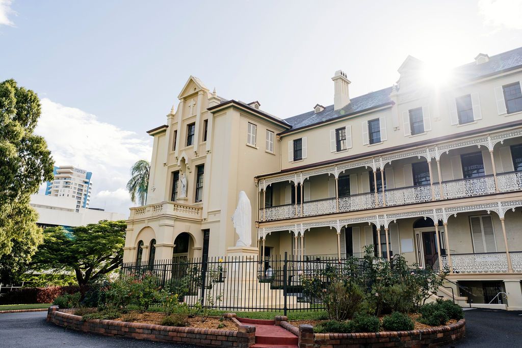 Historic two-story colonial building with wraparound verandas, ornate facade, and lush gardens in warm sunlight at All Hallows' School BNE