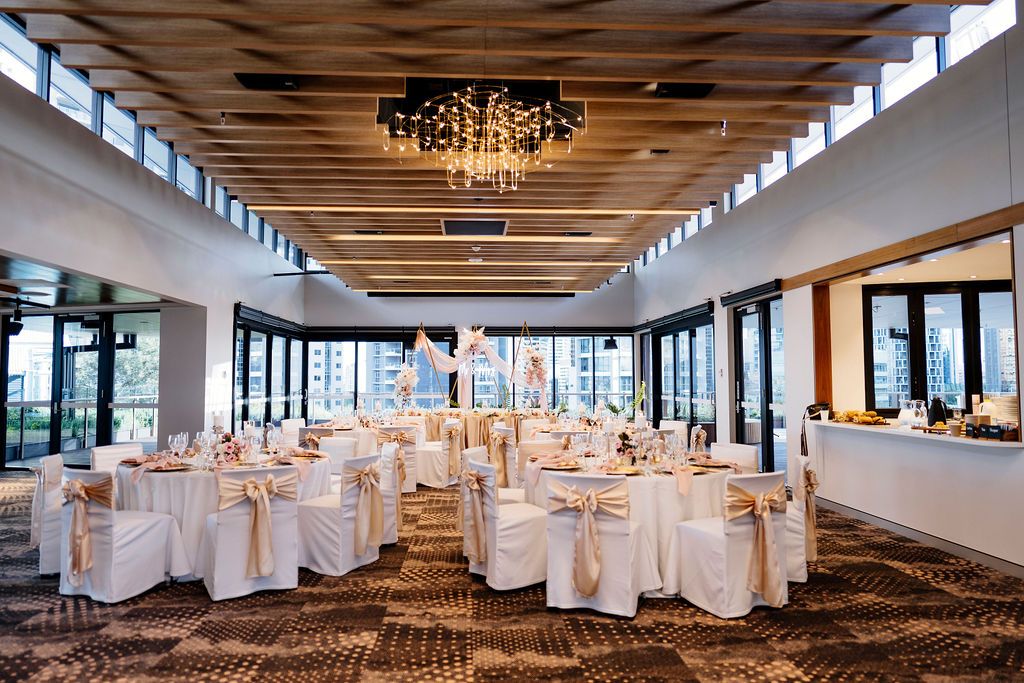 Elegant wedding reception hall with white chair covers, gold sashes, round tables, and a crystal chandelier All Hallows' School Brisbane