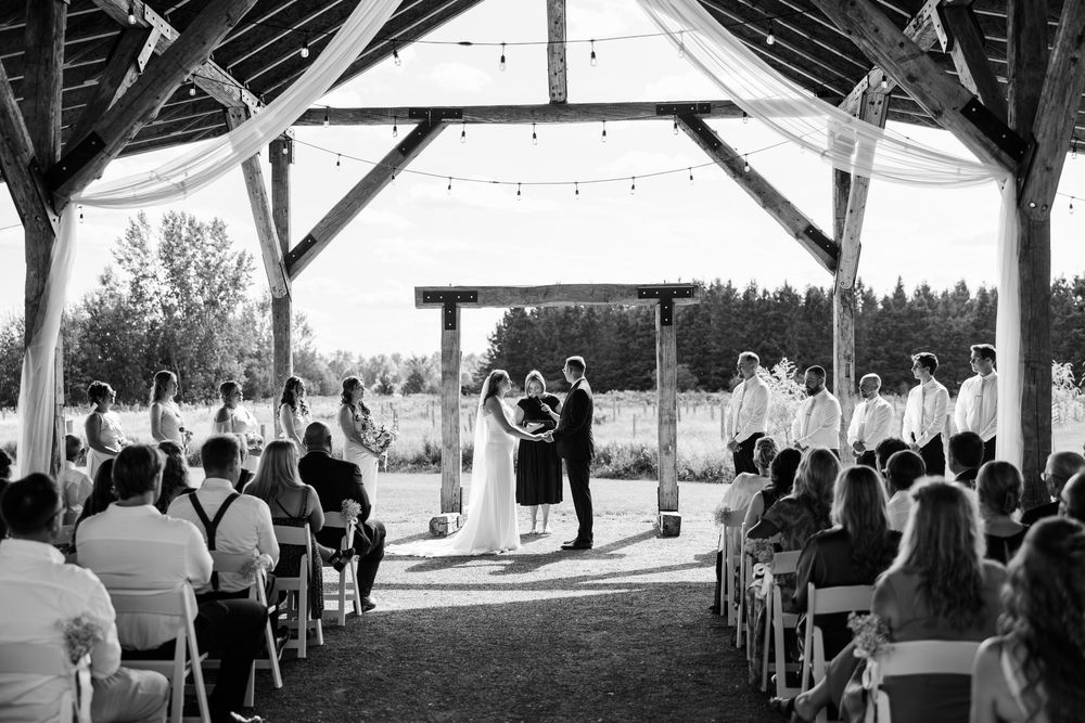 Black and white outdoor wedding ceremony under rustic wooden pavilion with string lights, guests seated in white chairs.