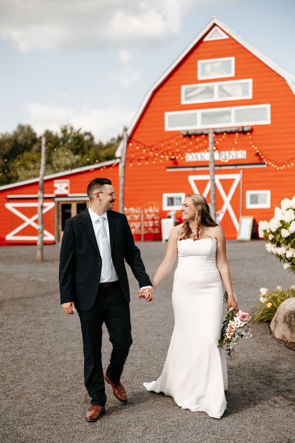 A bride and groom hold hands walking in front of a vibrant red barn decorated with string lights on their wedding day.