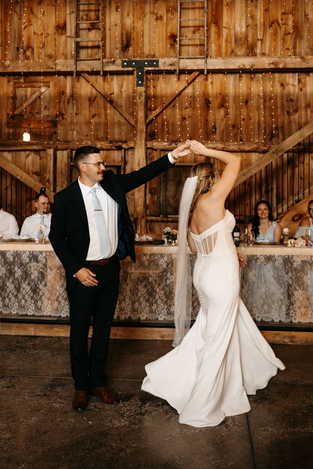 Bride and groom share their first dance in a rustic barn wedding venue with string lights and wooden walls.