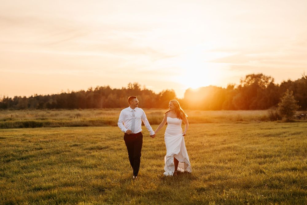 Bride and groom holding hands walking through a sunlit green field at golden hour sunset.