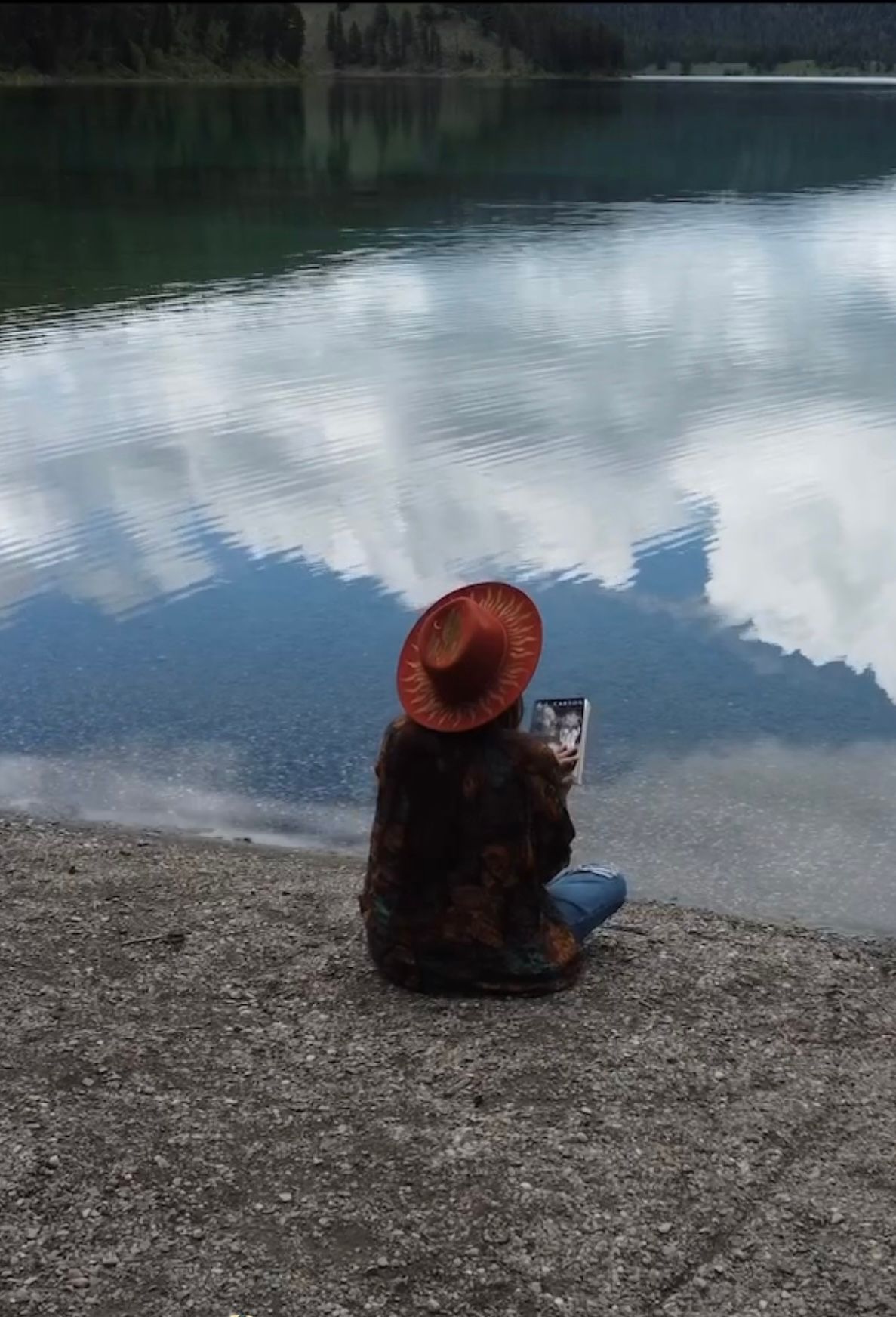 Person in red hat sitting lakeside reading, with stunning mountain reflections on calm water.