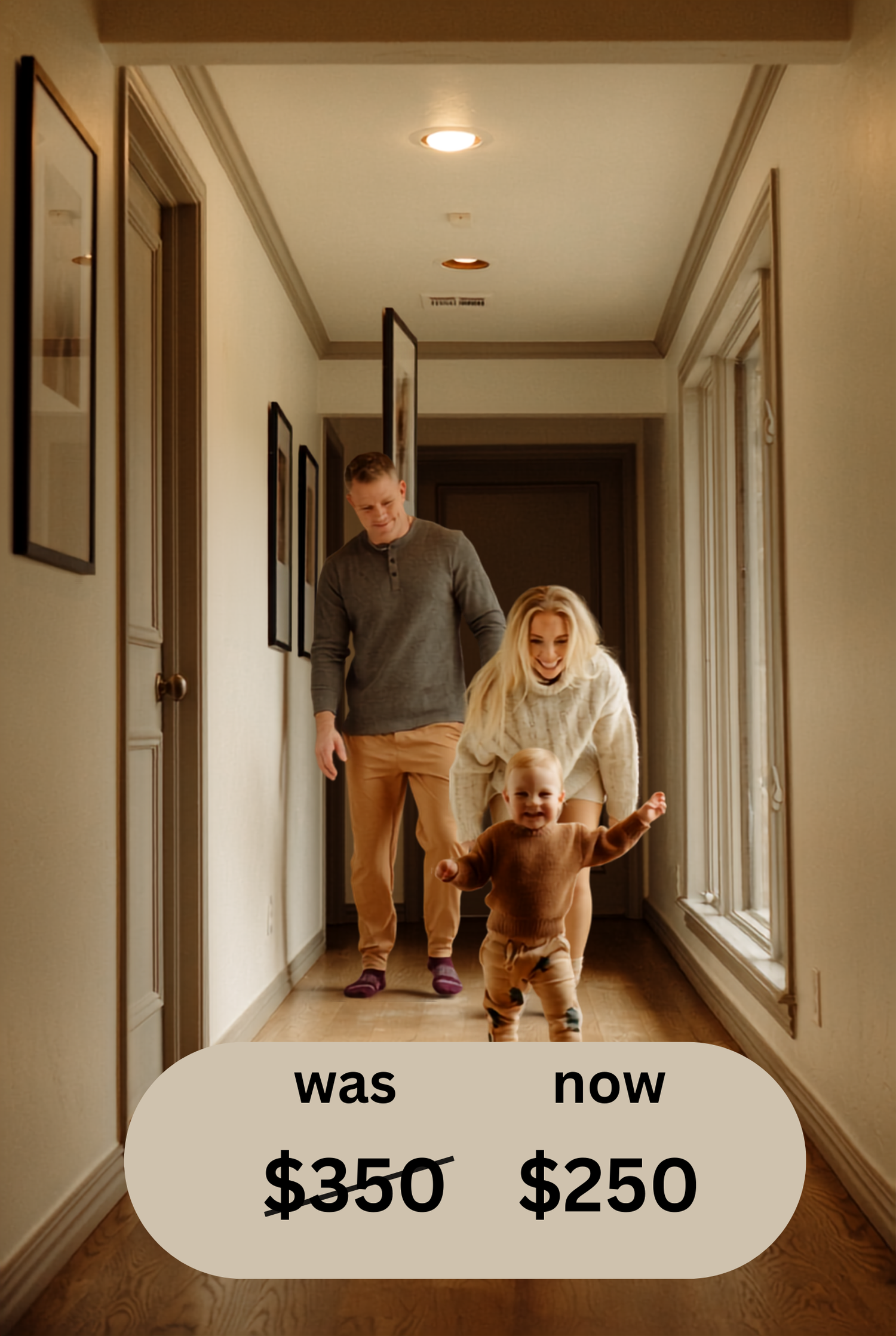 A toddler takes first steps in a hallway while parents watch proudly, framed photos on wall, warm wood floors.