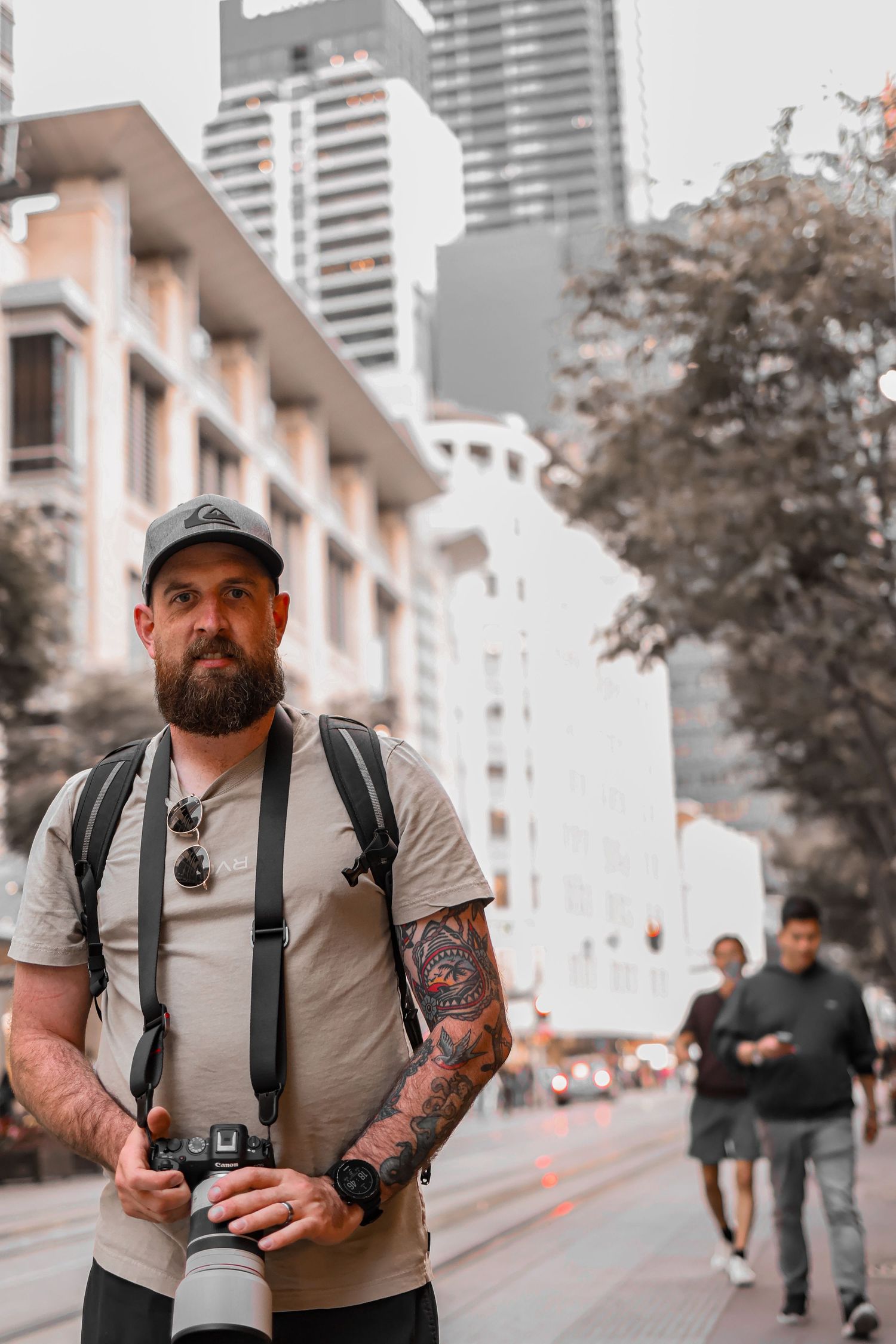 Tattooed photographer with cap and backpack holding a camera on a busy urban city street.