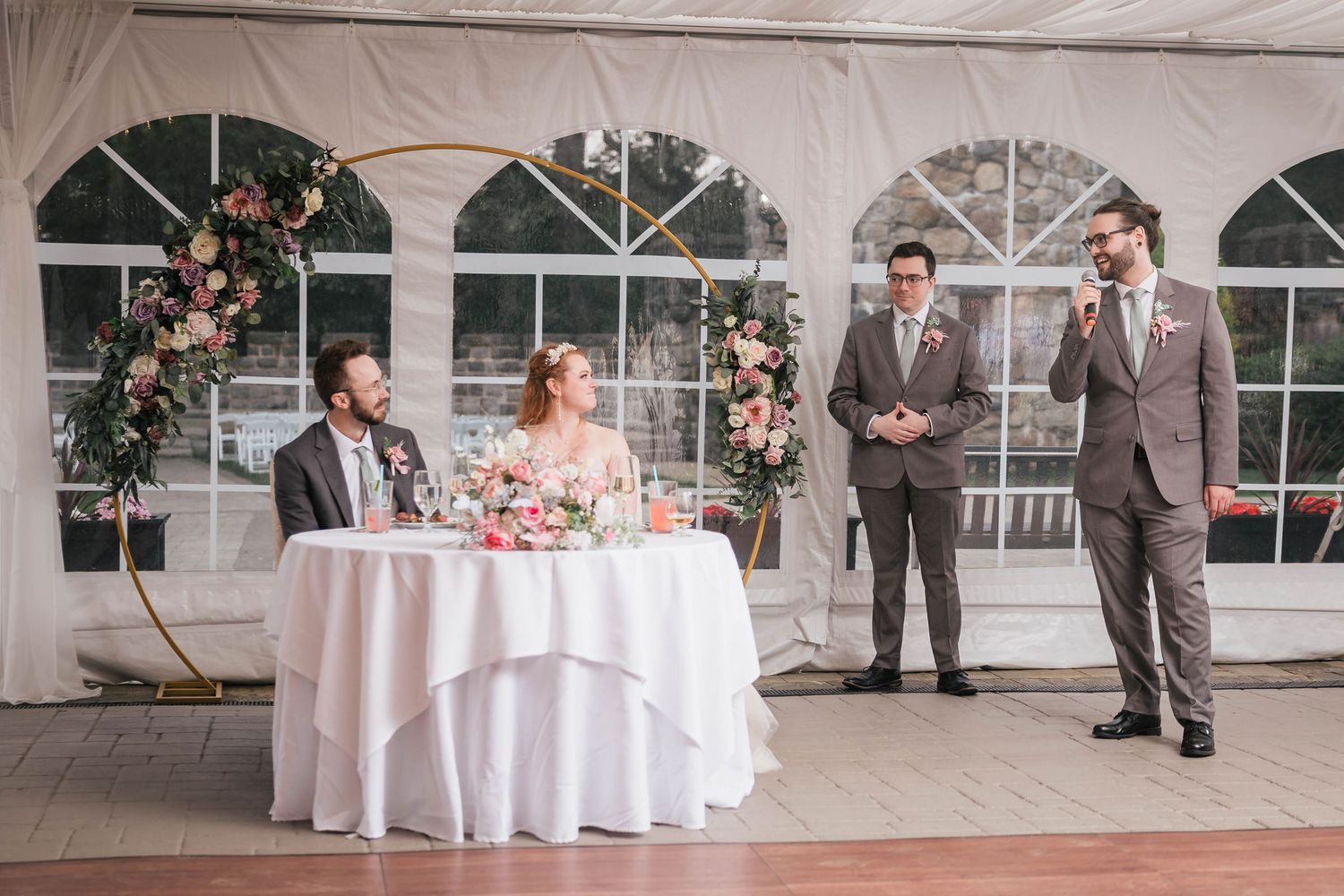 Groomsman giving a speech beside another groomsman, bride and groom listening at the sweetheart table.