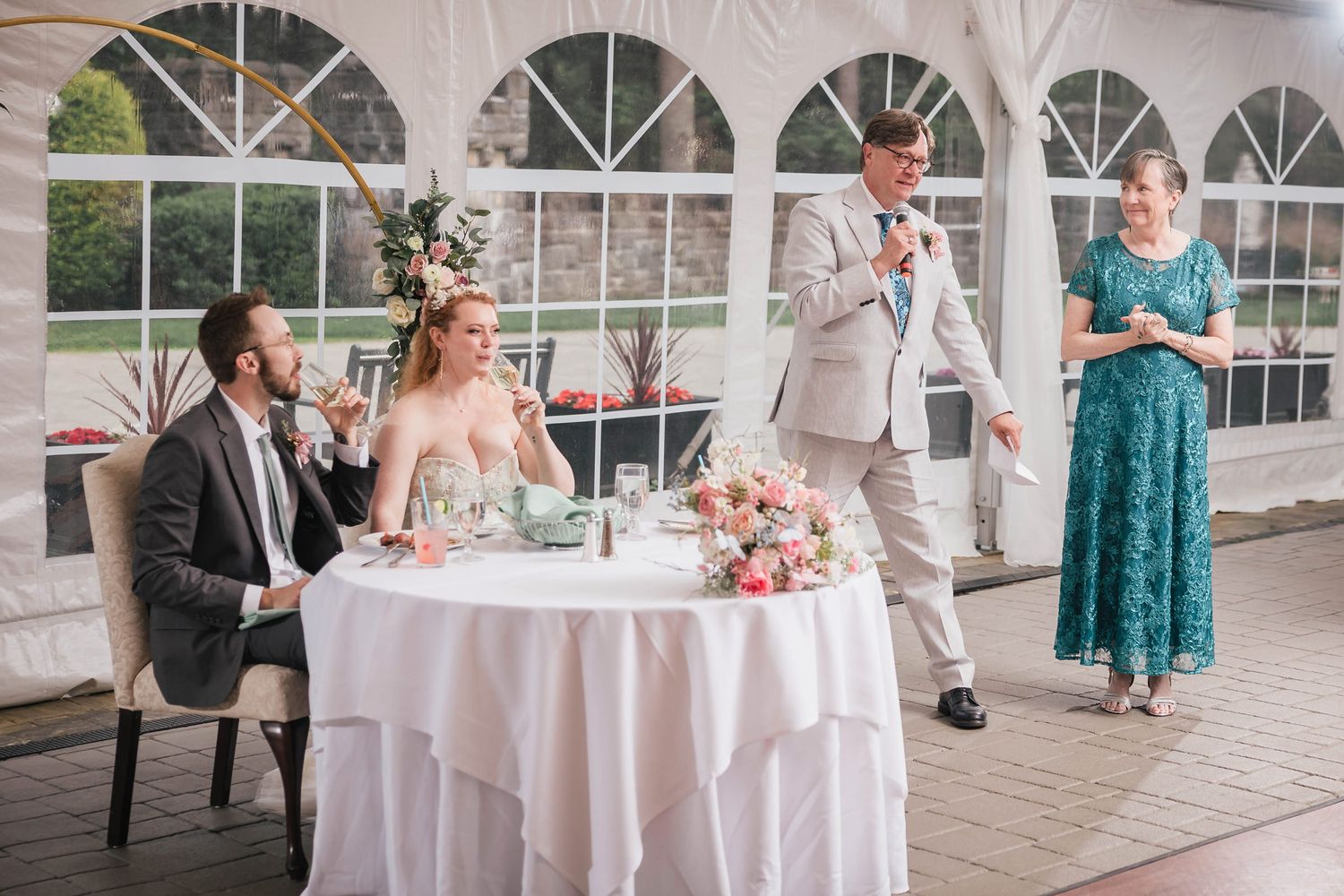 Groom's father mid-toast as the bride and groom raise their glasses to drink along with him.