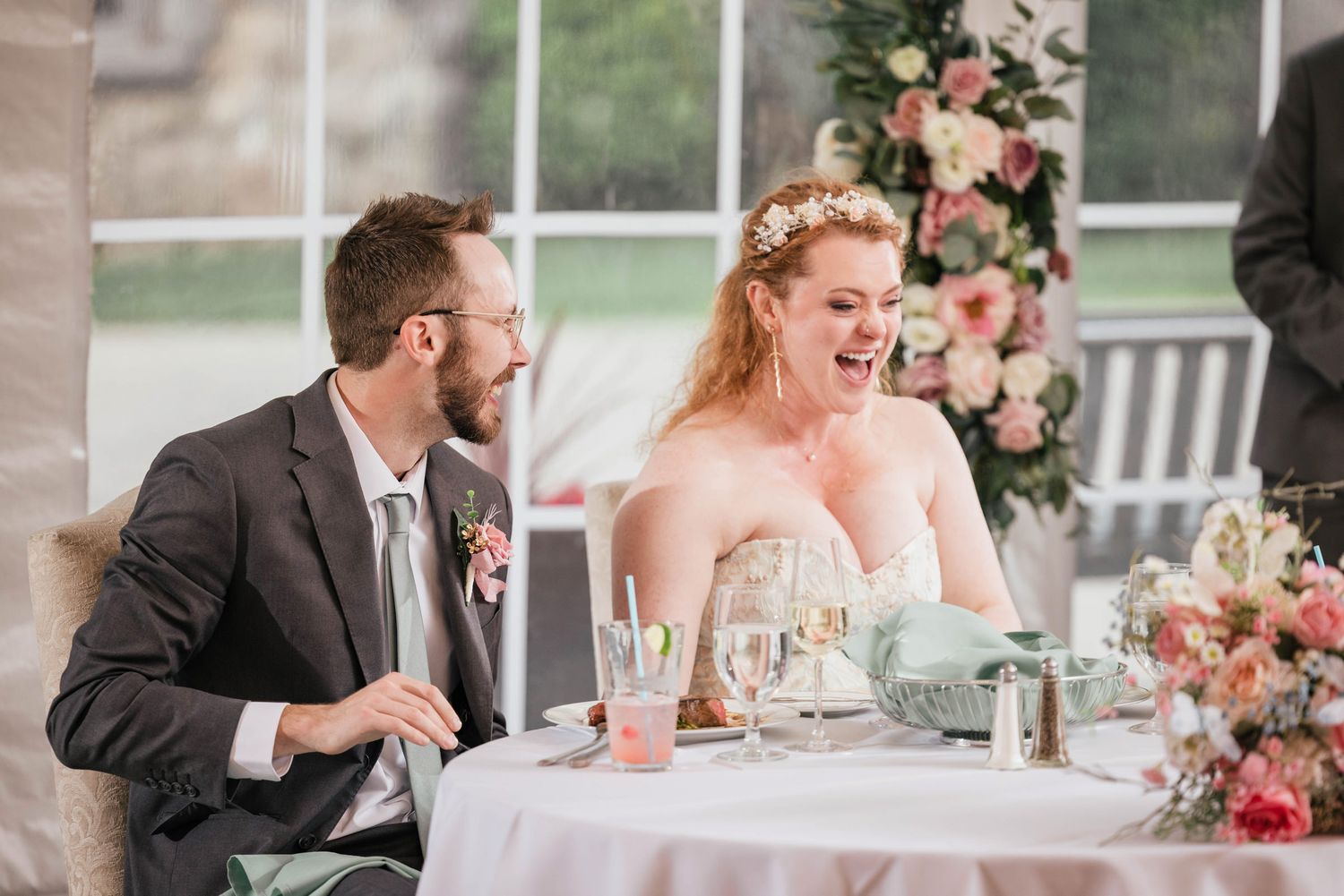 Bride and groom laughing together while listening to a speech at their table.