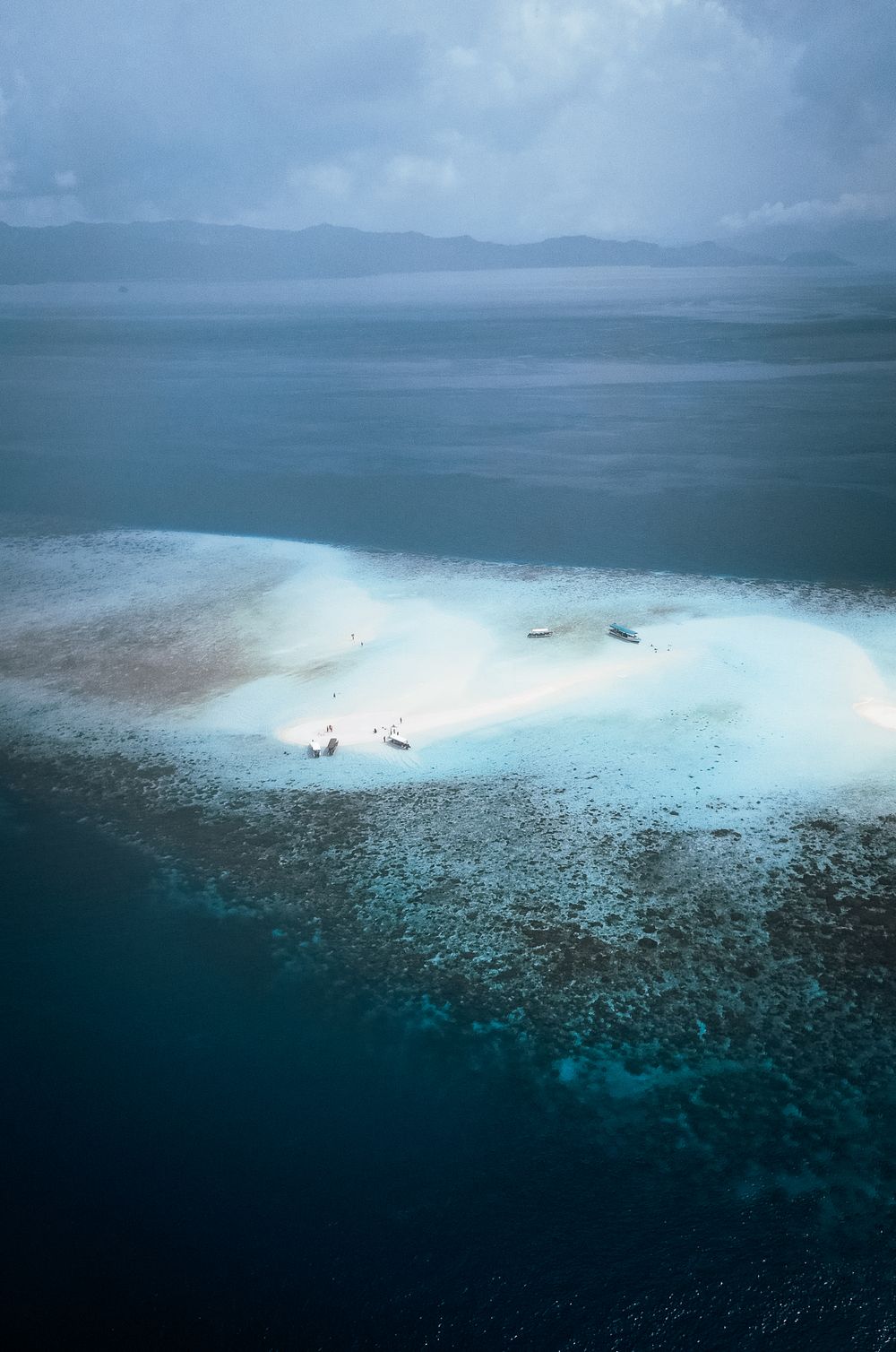 Aerial view of a tiny white sand island surrounded by coral reef and deep blue tropical ocean.