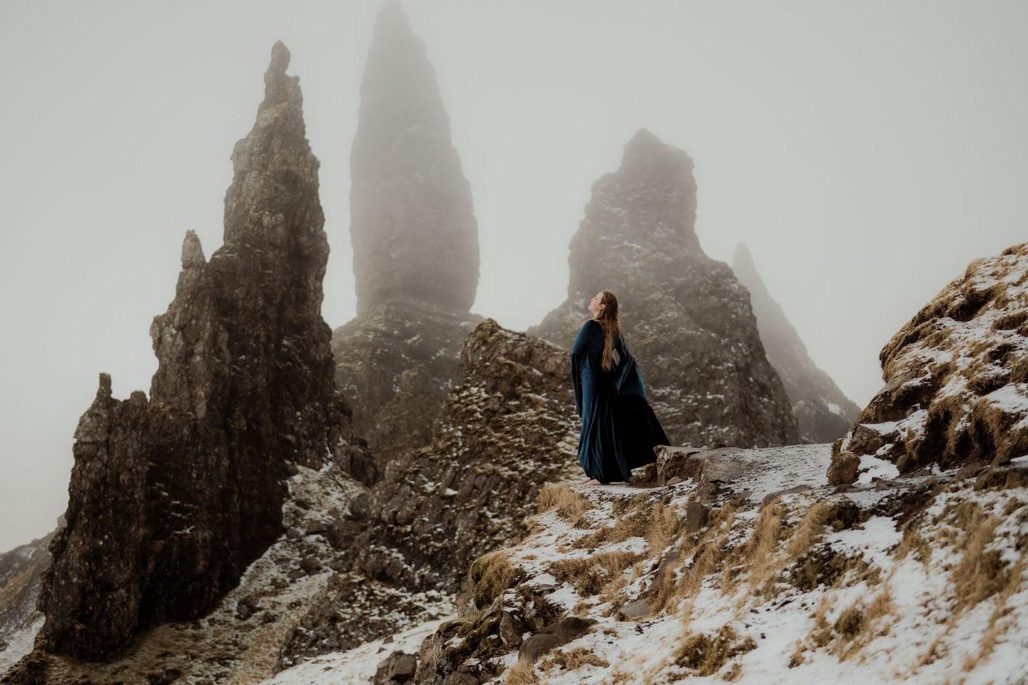Person in dark cloak standing among dramatic snow-dusted rock spires in misty Scottish Highlands landscape.