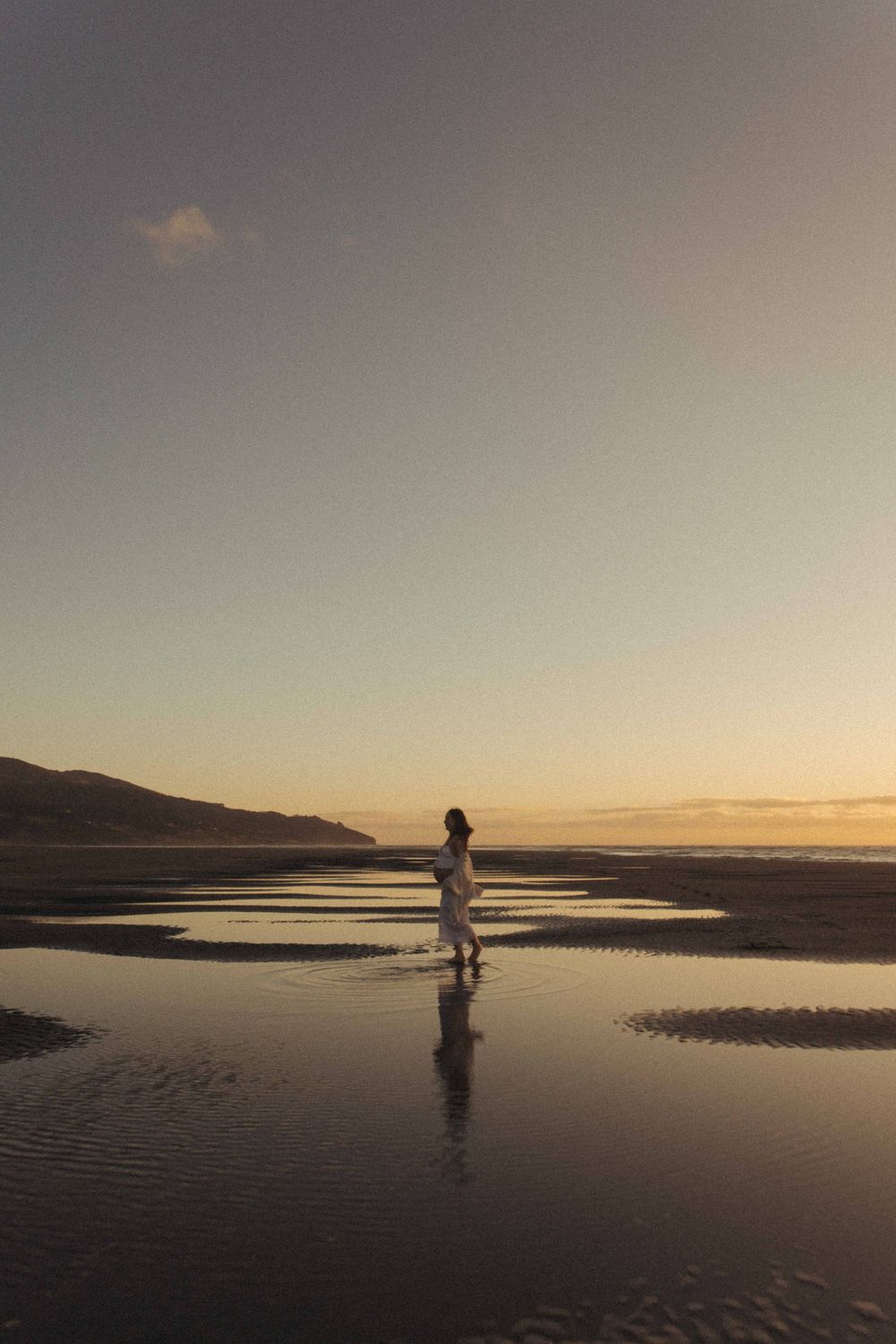 A pregnant woman in white walks on a reflective beach at golden sunset with hills in the background Raglan Waikato New Zealand.