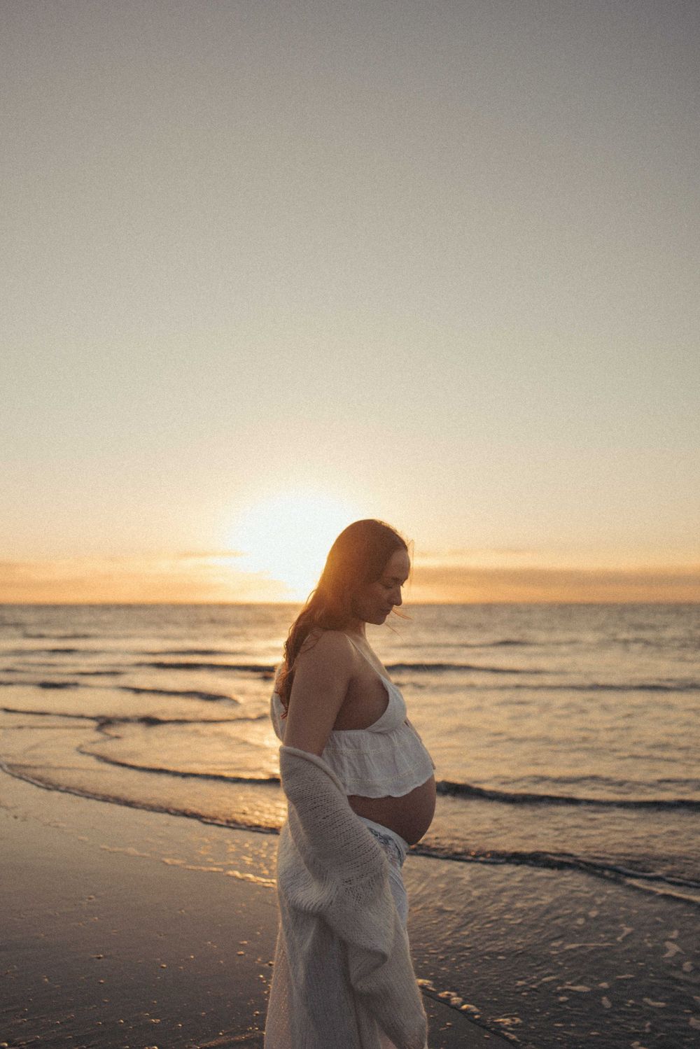 Pregnant woman in white dress cradling her baby bump on a serene beach at golden sunset in Raglan New Zealand.