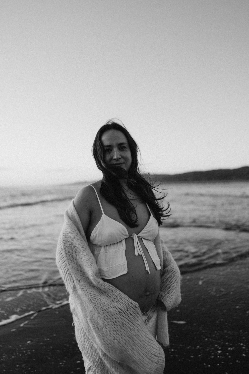 Black and white photo of a pregnant woman in a bikini and sheer wrap standing on a beach at dusk in Raglan New Zealand.