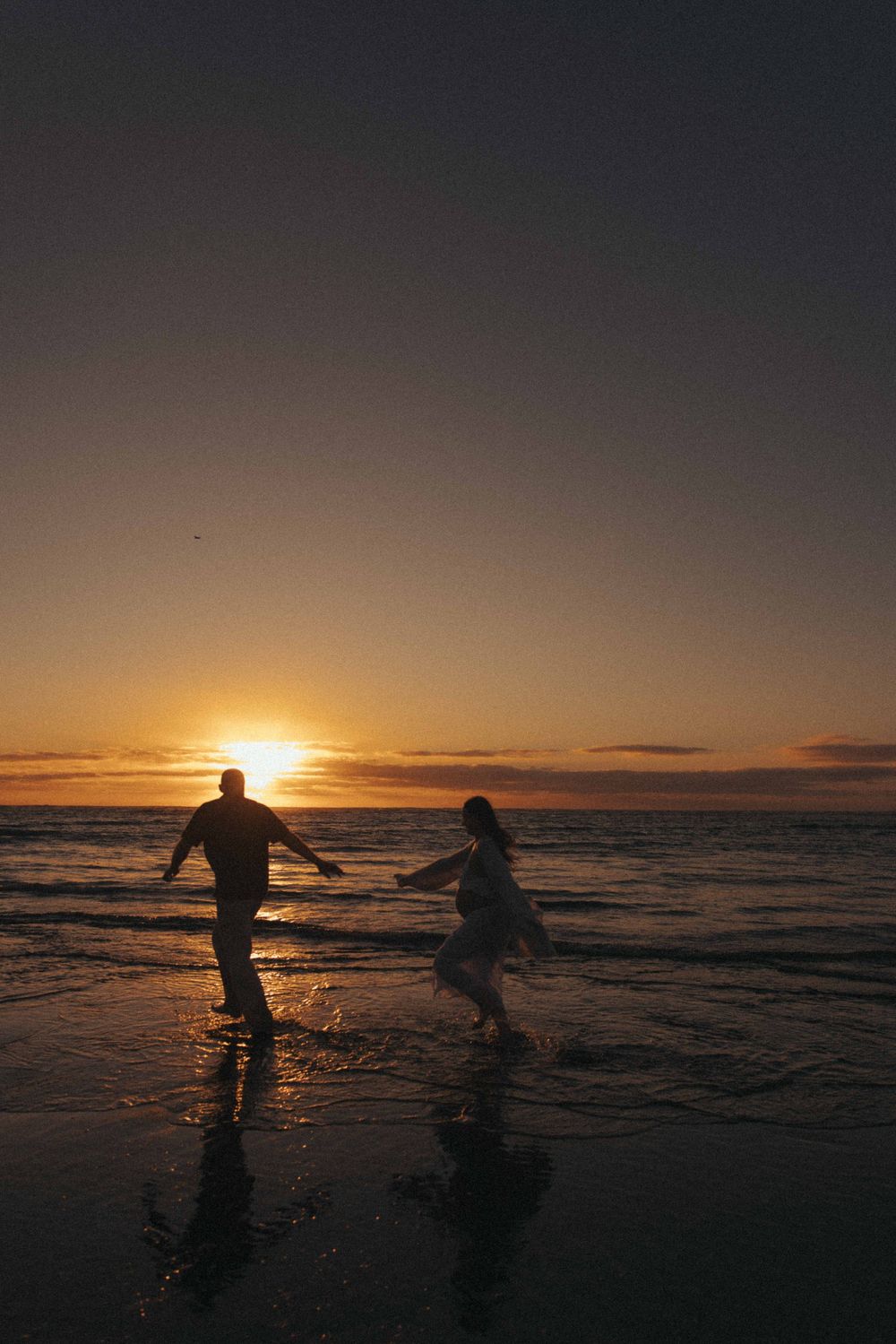 Couple running together on a beach at sunset, silhouetted against golden ocean waves and vibrant sky in Raglan New Zealand.