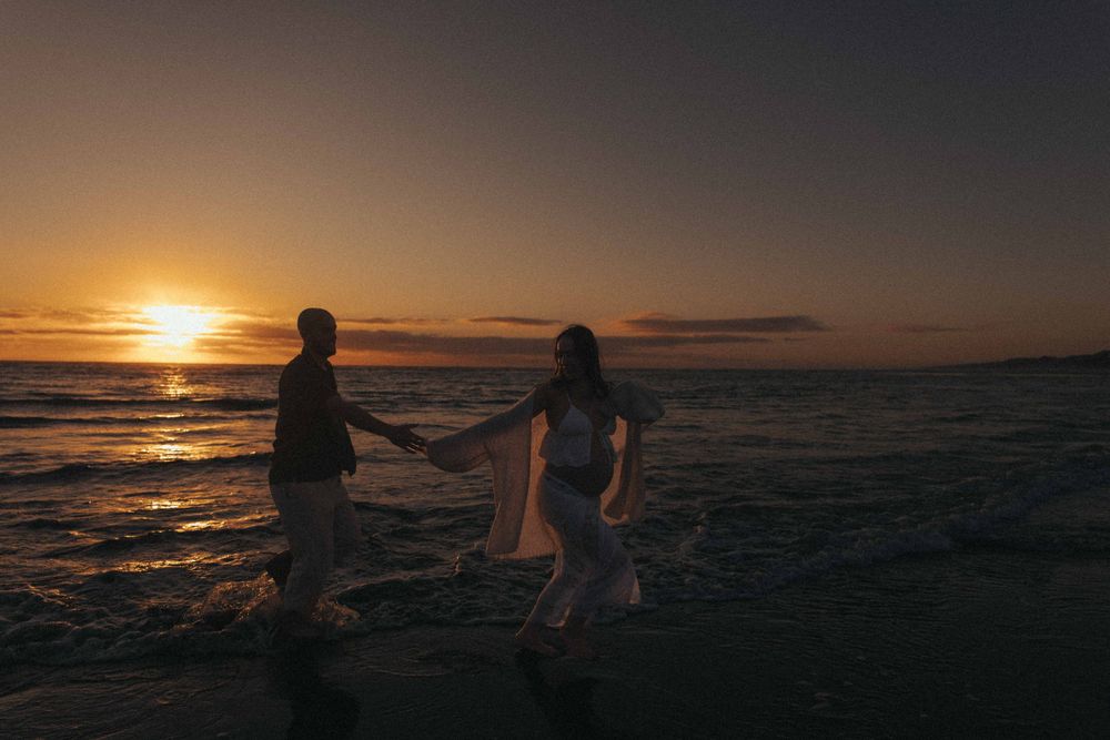 Couple holding hands walking on a sandy beach at sunset with golden sky and ocean waves in Raglan New Zealand.