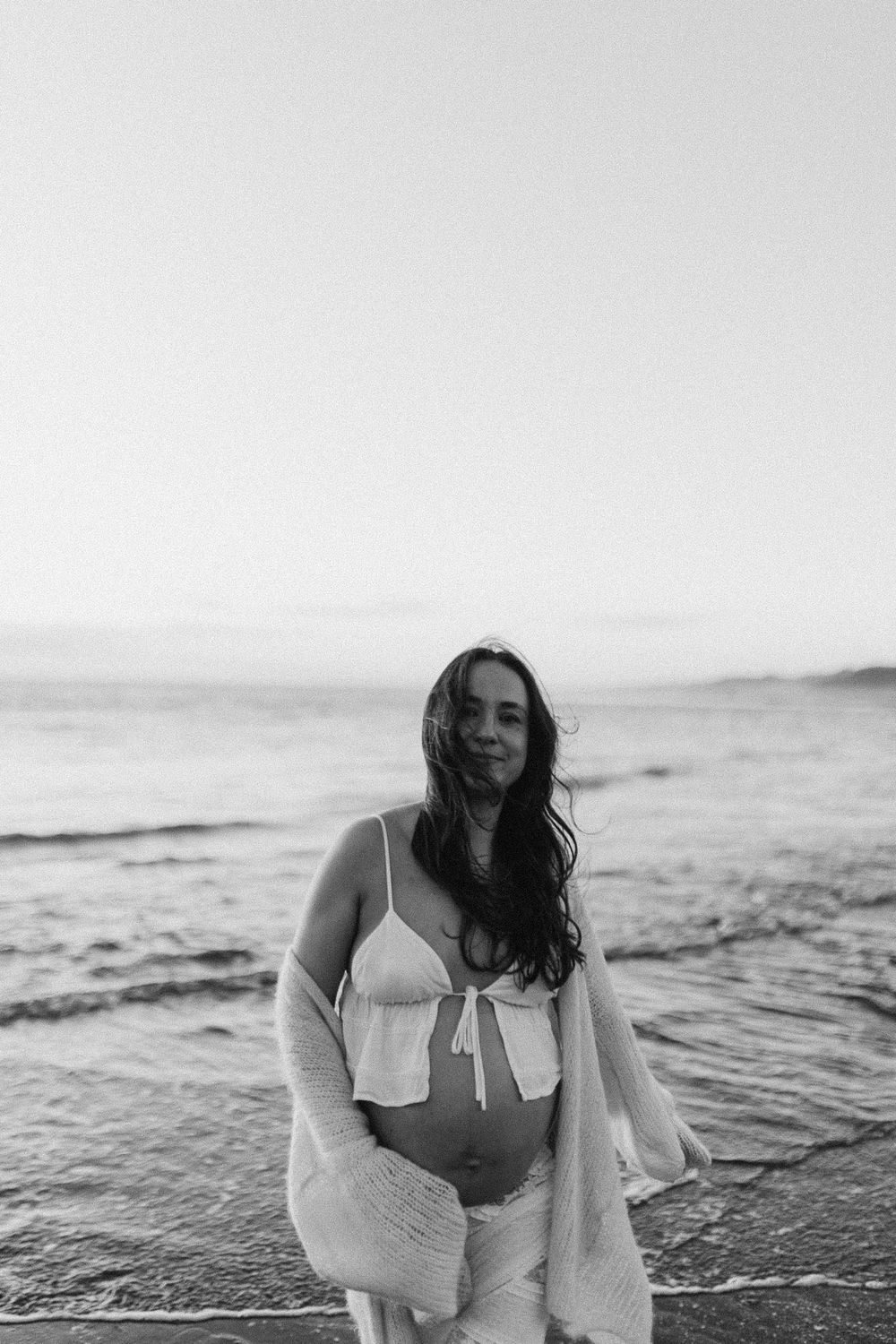 Black and white photo of a pregnant woman in a bikini top and cardigan standing on a beach at dusk in Raglan New Zealand.