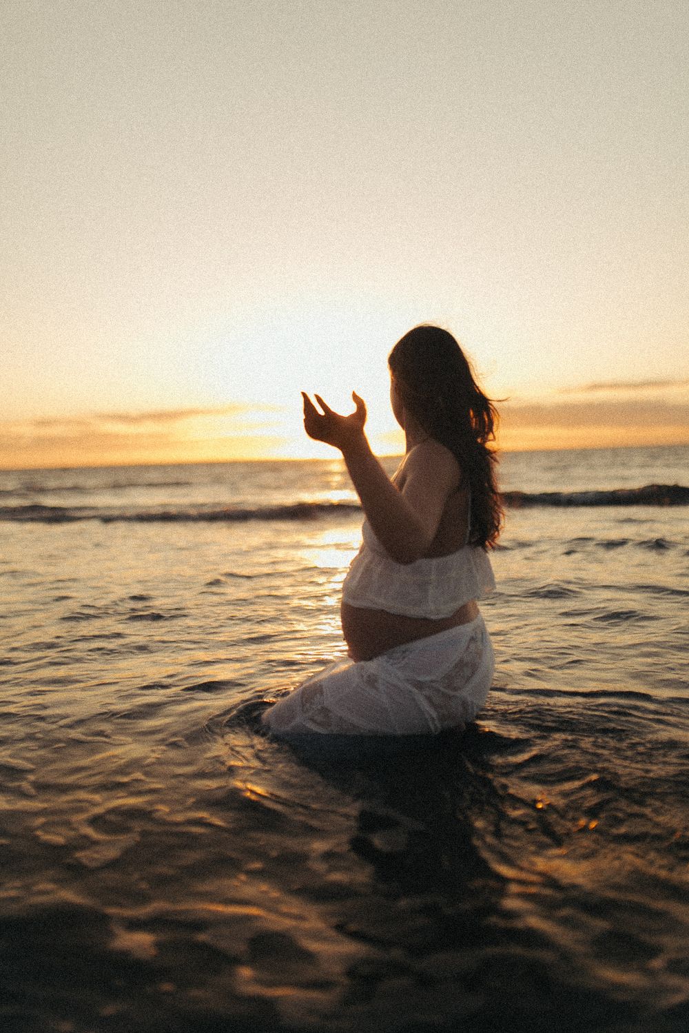 Pregnant woman in white dress standing in ocean waves during golden sunset, holding hand up gracefully in Raglan New Zealand.