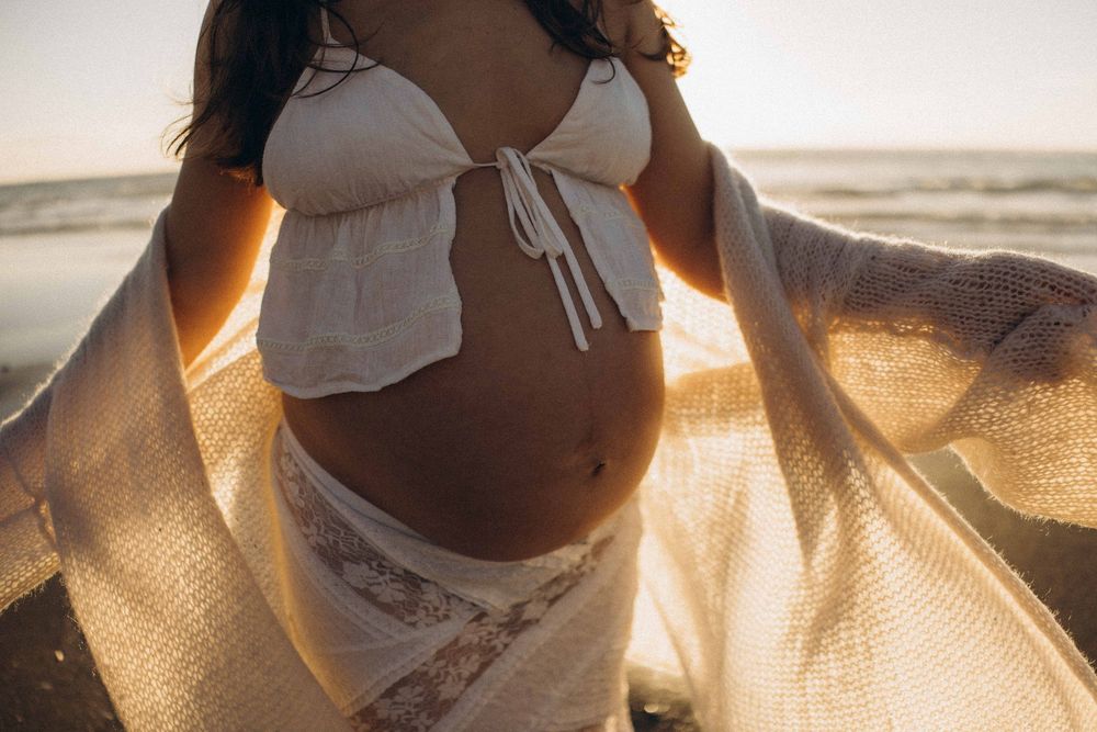 Pregnant woman in white bikini top and lace skirt posing on beach at golden sunset, outdoor maternity photography on Raglan beach