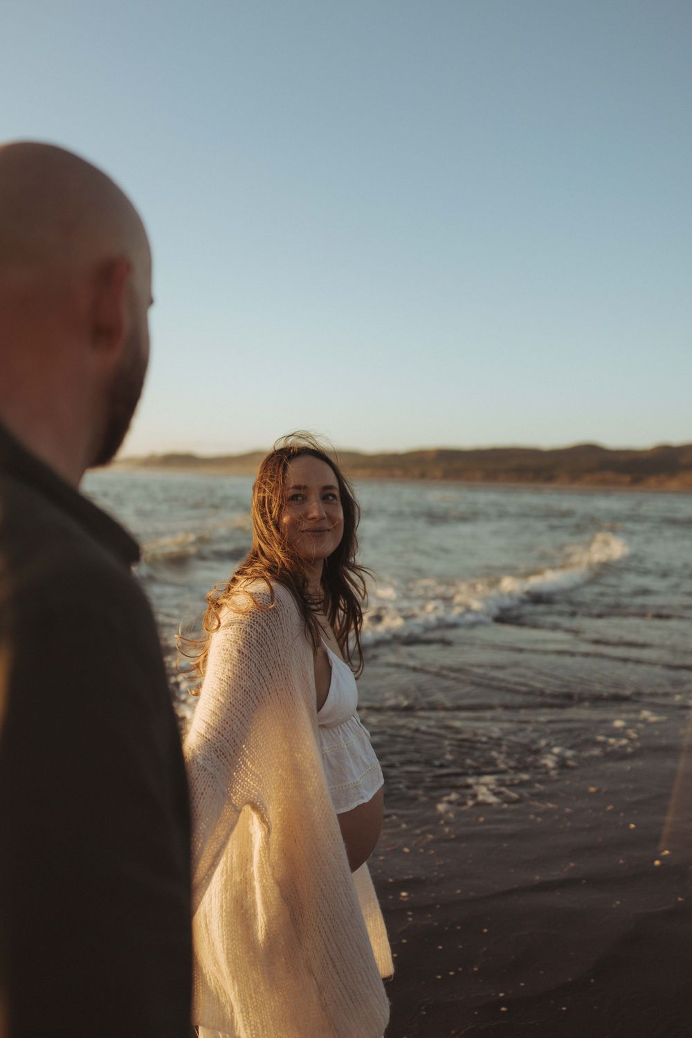 Woman in white dress smiling at man on beach during golden hour sunset maternity couple photography New Zealand.