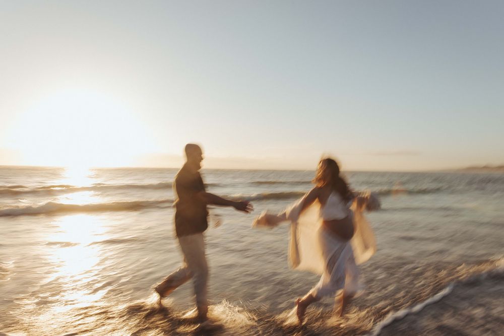 Couple holding hands running along a sandy beach at golden sunset with ocean waves in Raglan New Zealand.